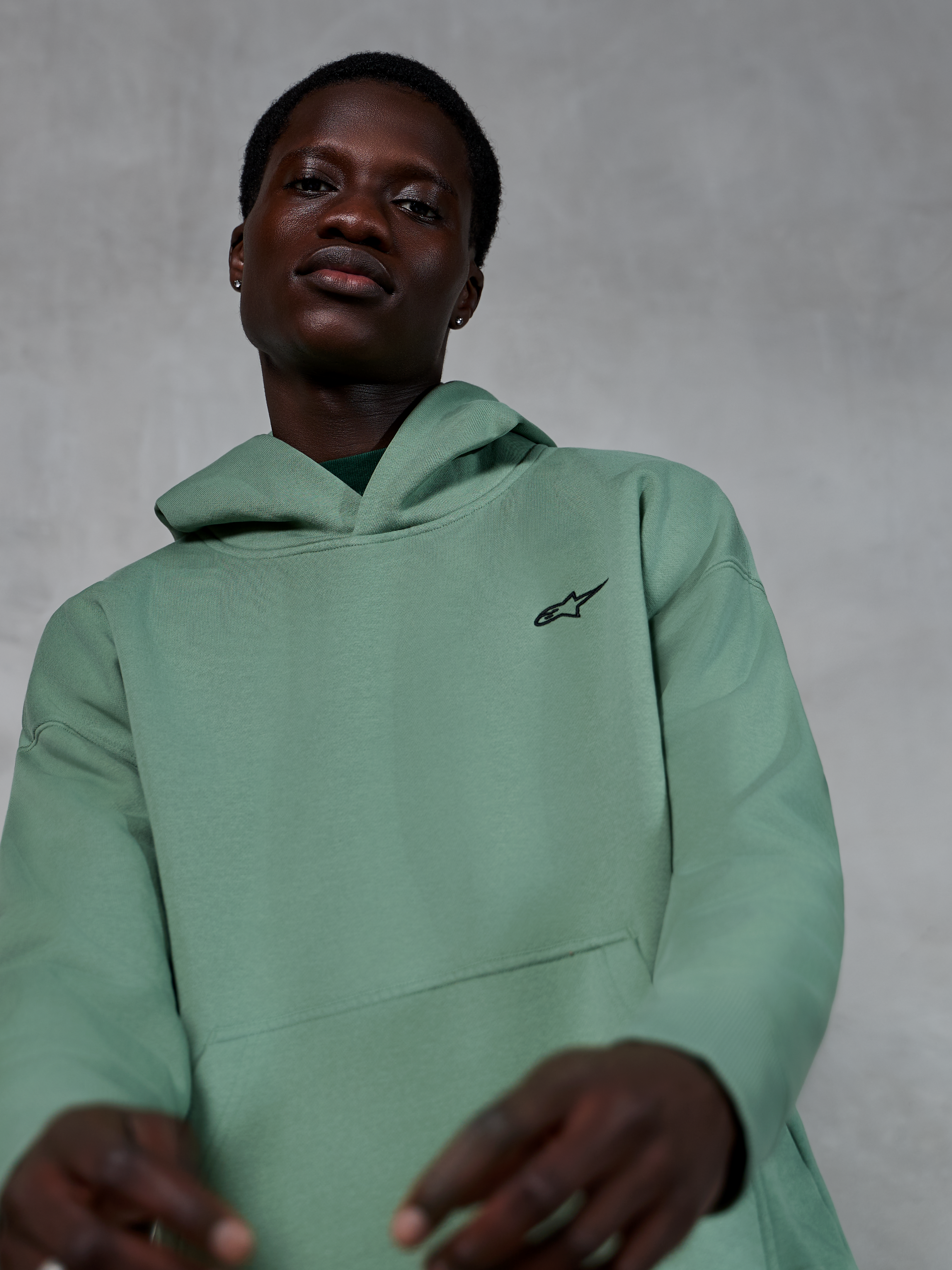 Felpa con cappuccio Alpinestars Essentials Ageless, abbigliamento sportivo casual in verde Cactus, indossata da un modello, con un piccolo logo Alpinestars nero sul petto, pile di alta qualità con vestibilità rilassata ed estetica pulita