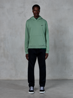 Felpa con cappuccio Alpinestars Essentials Ageless, verde Cactus, piccolo logo nero sul petto, tasca a marsupio, streetwear casual indossato con pantaloni neri e sneaker bianche e nere contro una parete strutturata
