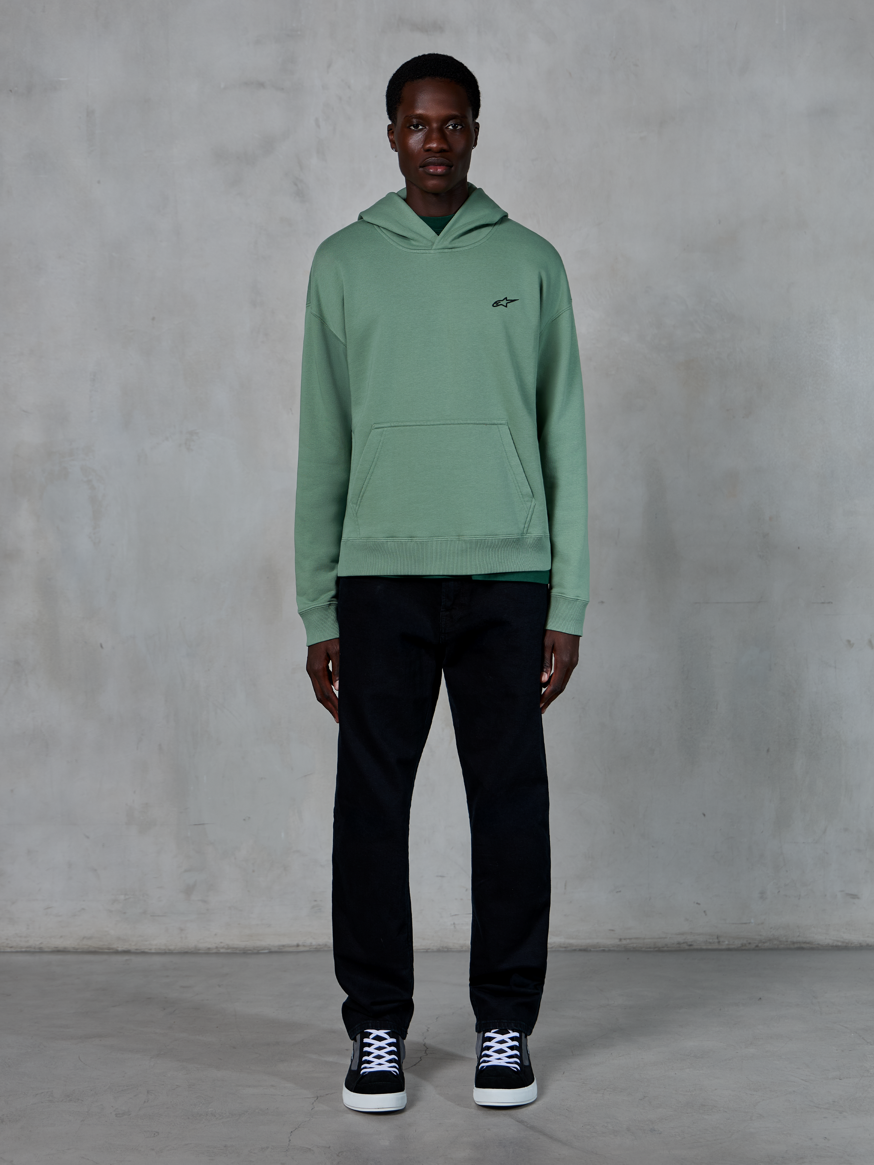 Felpa con cappuccio Alpinestars Essentials Ageless, verde Cactus, piccolo logo nero sul petto, tasca a marsupio, streetwear casual indossato con pantaloni neri e sneaker bianche e nere contro una parete strutturata