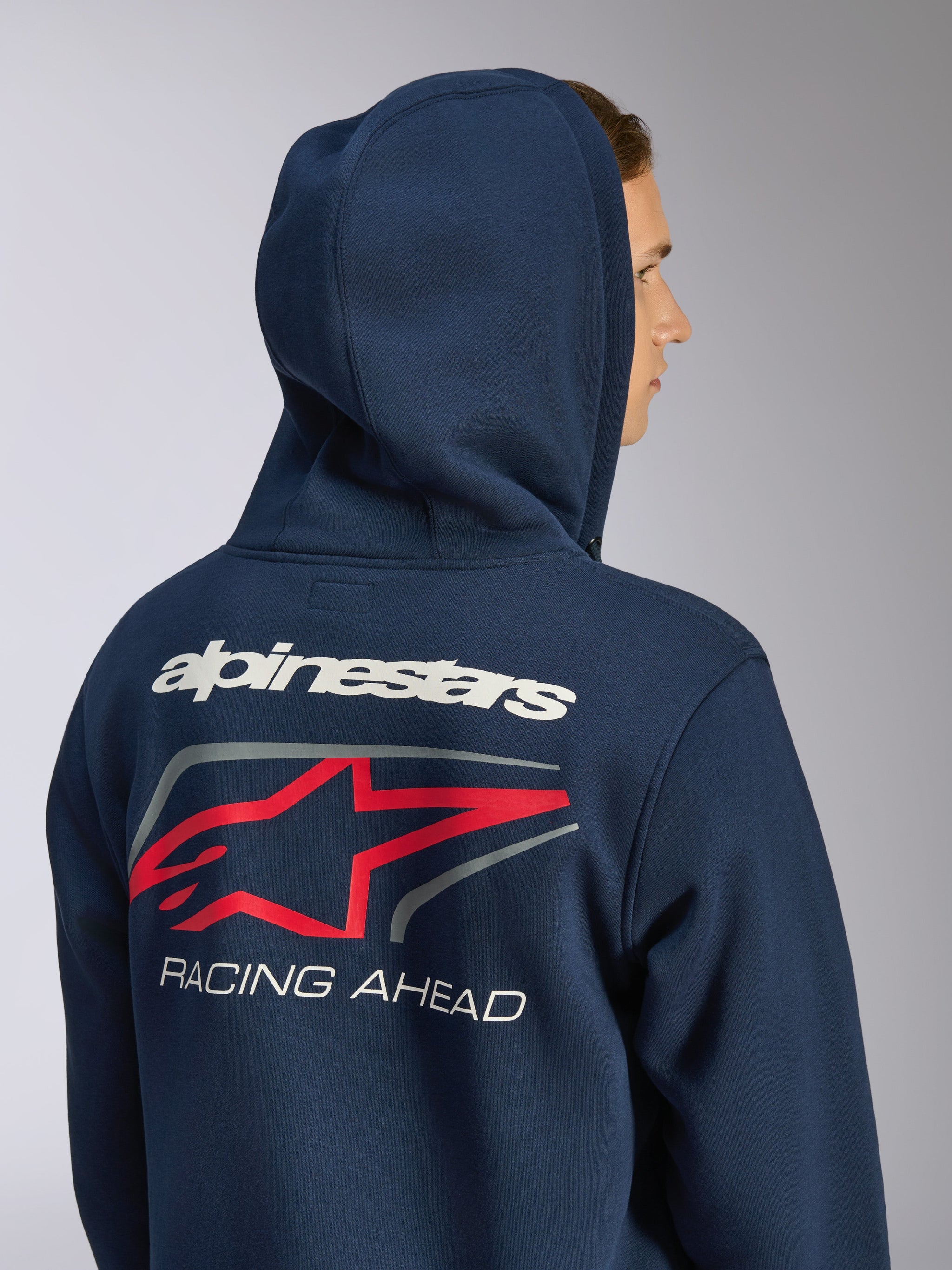 Felpa con cappuccio Formulation Alpinestars, pile blu navy con zip frontale, vista posteriore con cappuccio sollevato e grande grafica posteriore con marchio Alpinestars bianco, logo a stella rosso e scritta 'RACING AHEAD', streetwear casual ispirato al motorsport