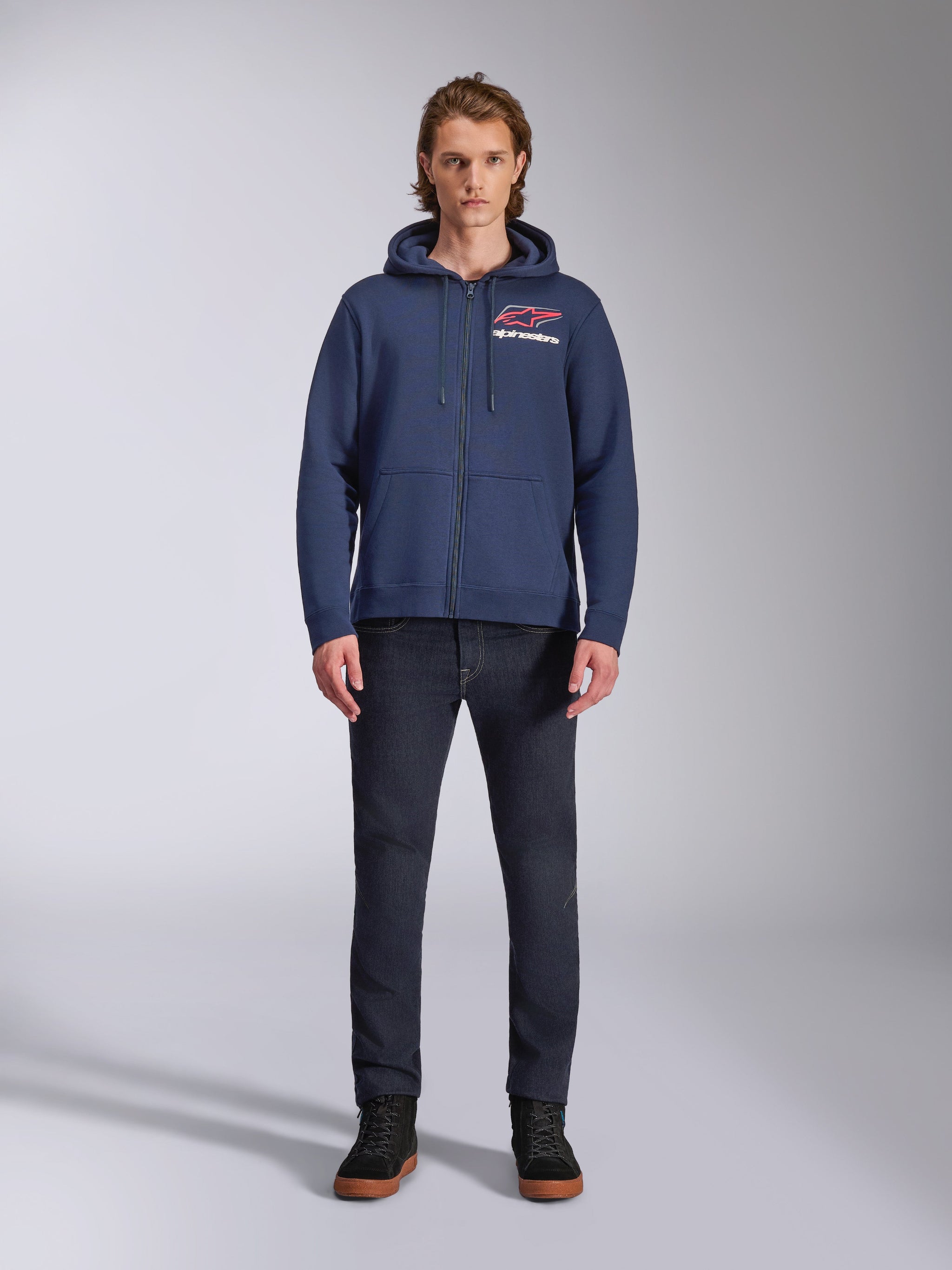 Felpa con cappuccio Alpinestars Formulation, felpa in pile blu navy con zip, indossata da un modello con jeans blu scuro, logo rosso e bianco sul petto, cappuccio foderato in rete con cordini, streetwear casual motorsport