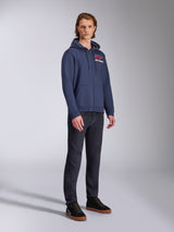 Modello che indossa la felpa con cappuccio Alpinestars Formulation, pile in poliestere blu navy con zip frontale, logo rosso e bianco sul petto, abbinata a jeans scuri e sneakers nere, abbigliamento streetwear casual ispirato al motorsport