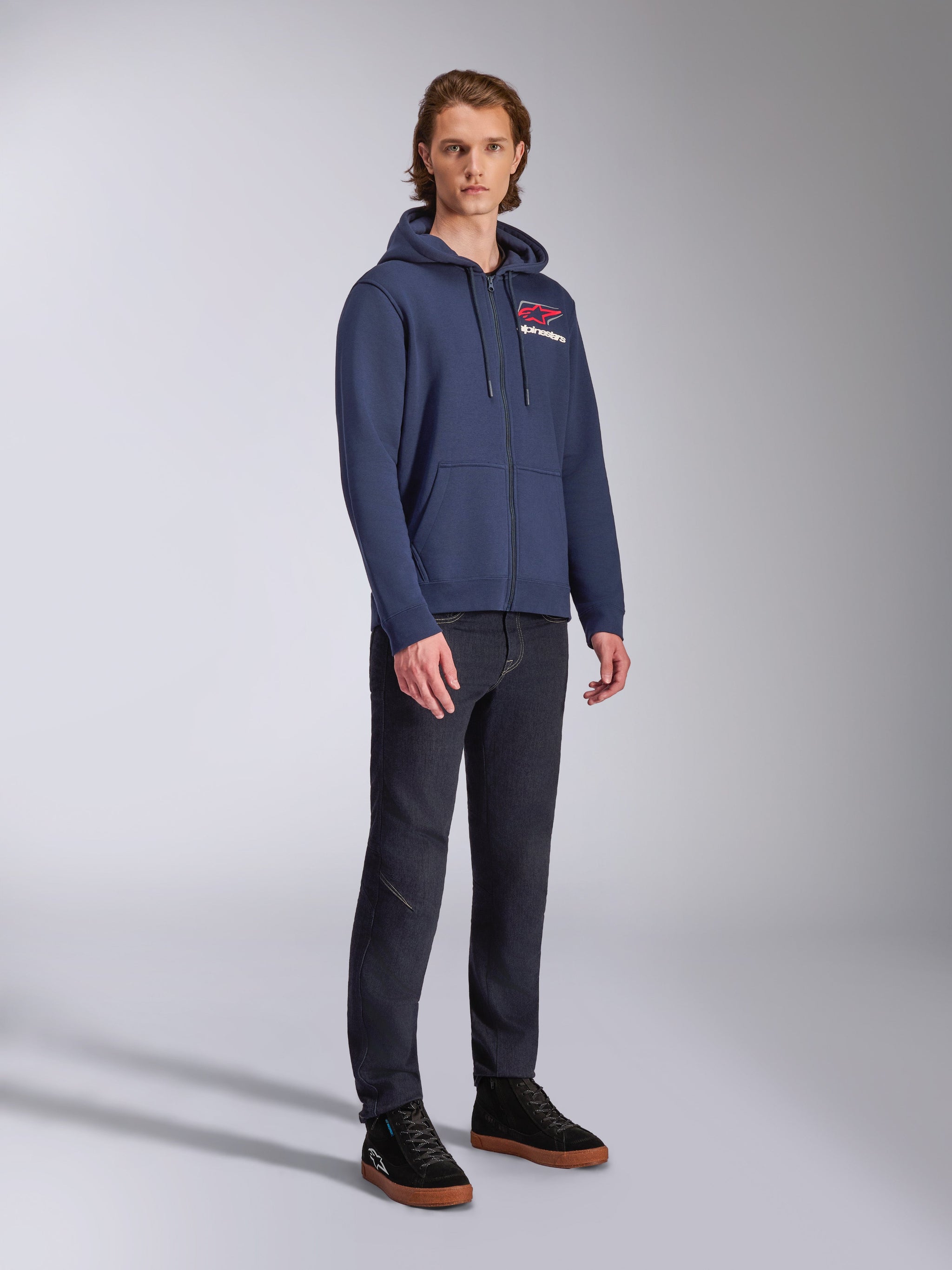 Modello che indossa la felpa con cappuccio Alpinestars Formulation, pile in poliestere blu navy con zip frontale, logo rosso e bianco sul petto, abbinata a jeans scuri e sneakers nere, abbigliamento streetwear casual ispirato al motorsport