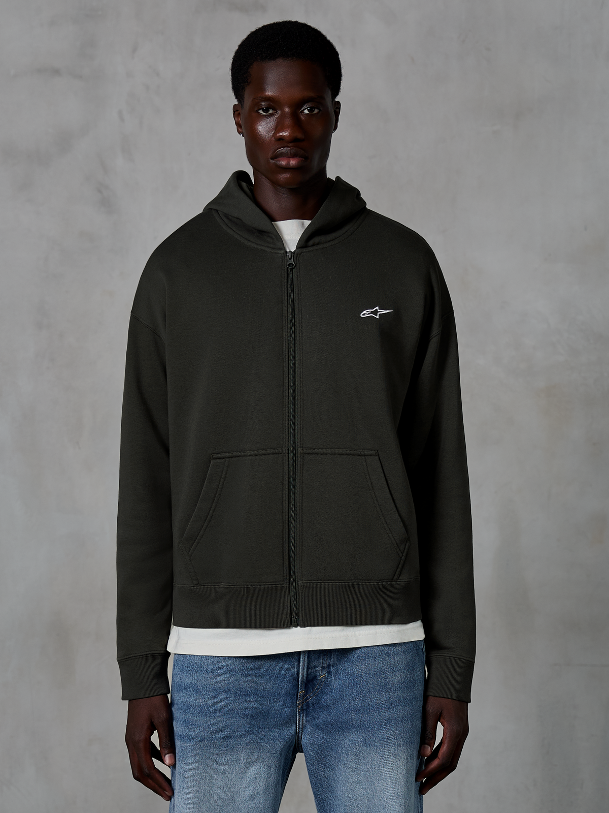 Alpinestars Essentials Ageless Zip Hoodie, felpa con cappuccio e zip integrale, Off Black, con un piccolo logo a stella bianco sul petto, tasche frontali e polsini a costine, indossata come abbigliamento streetwear casual ispirato al motorsport