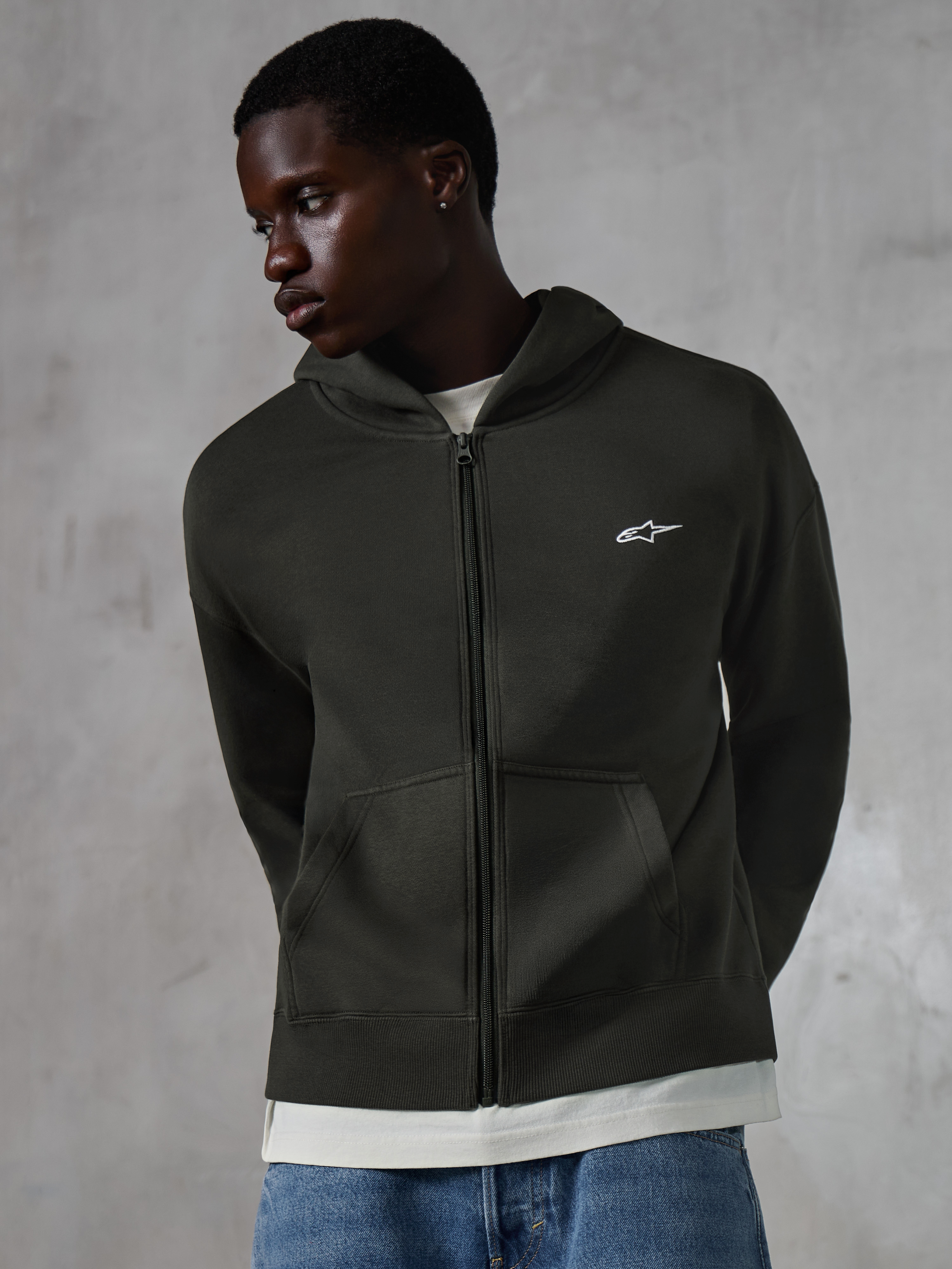Alpinestars Essentials Ageless Zip Hoodie, streetwear casual Off Black, felpa con cappuccio e zip integrale con piccolo logo Alpinestars bianco sul petto, indossata da un modello con jeans blu, vestibilità rilassata, tasche anteriori e finitura stone-washed.