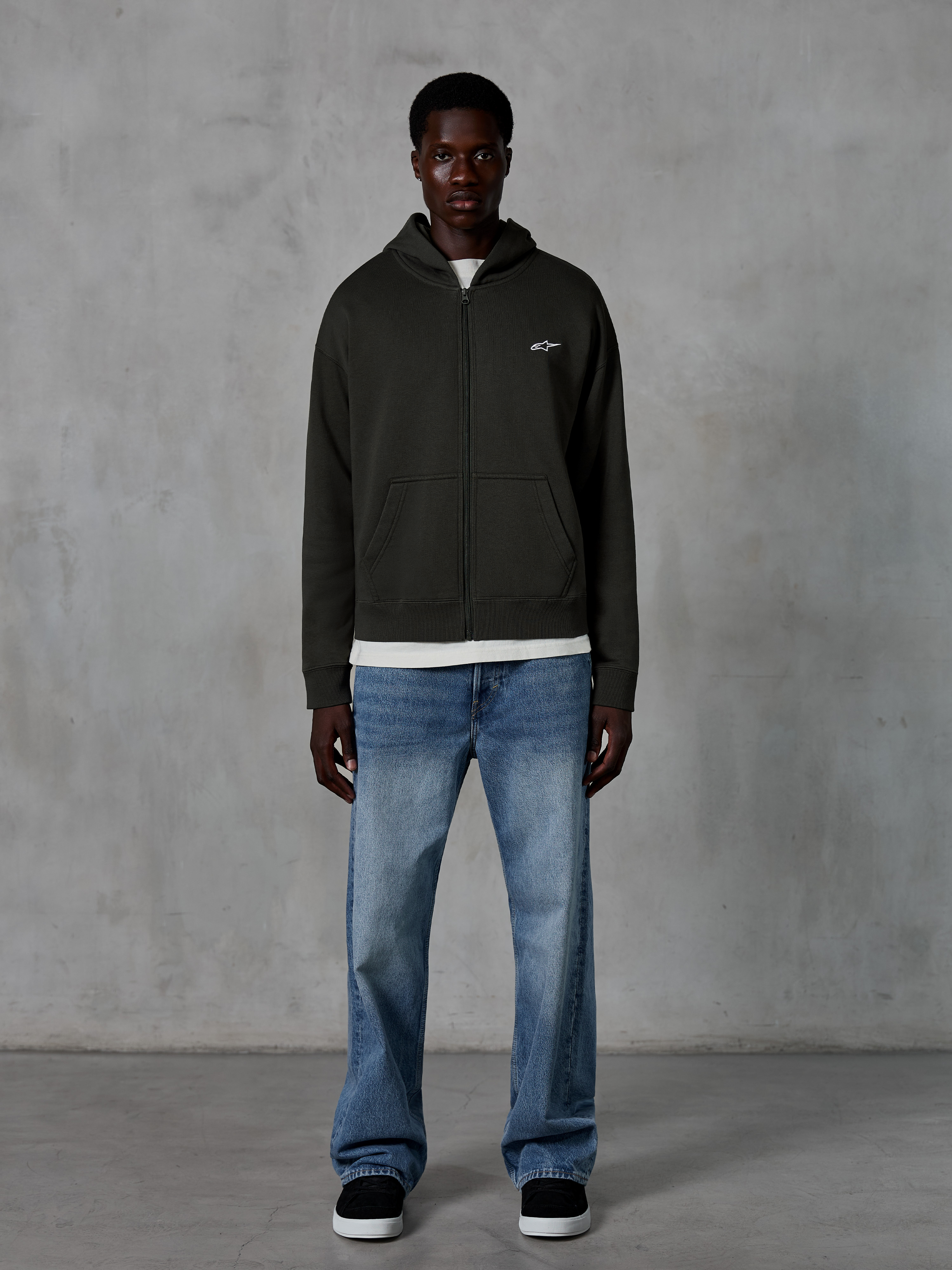 Alpinestars Essentials Ageless Zip Hoodie, felpa con cappuccio e zip integrale, Off Black, con un piccolo logo Alpinestars bianco sul petto, indossata da un modello con t-shirt bianca e jeans blu, vestibilità rilassata, streetwear casual ispirato al motorsport