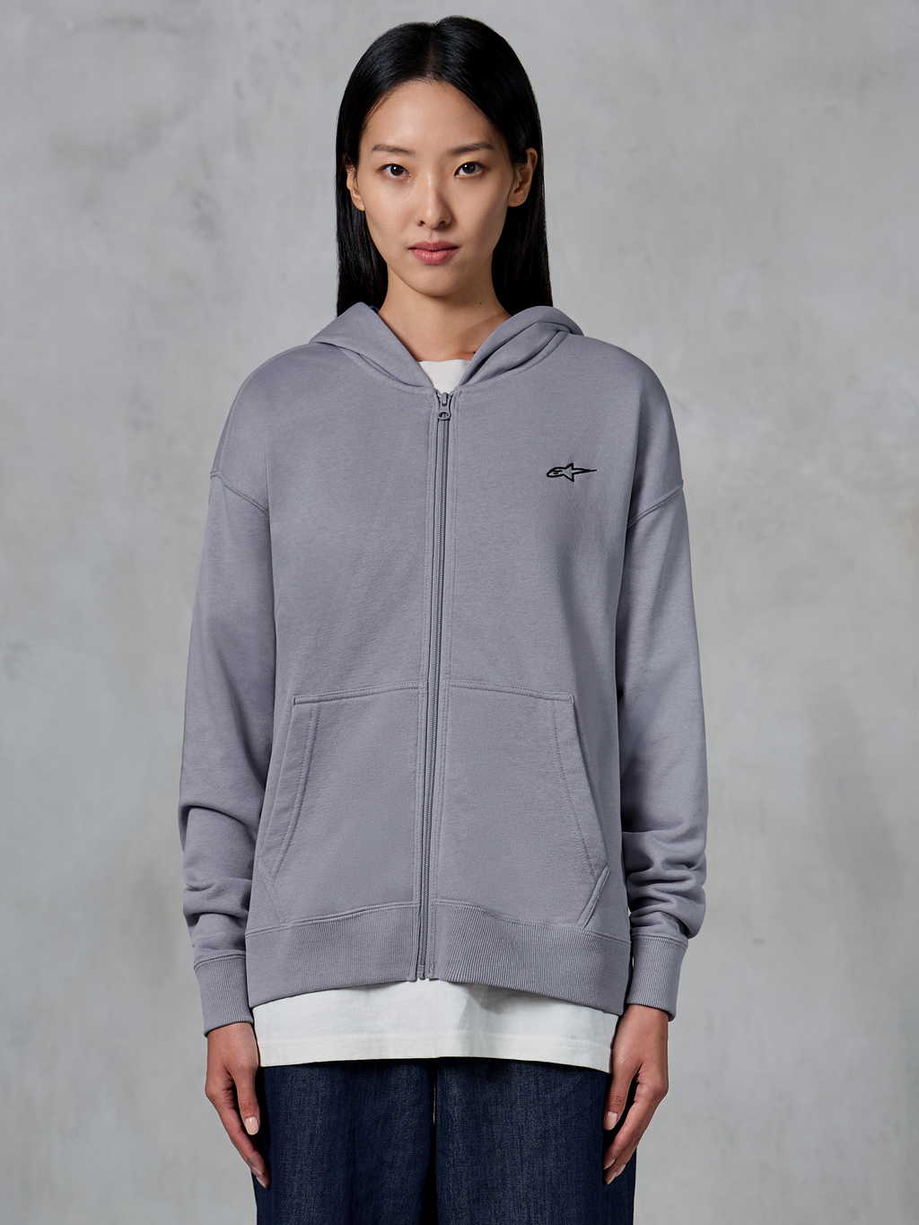 Felpa con cappuccio Alpinestars Essentials Ageless Zip, Silver, streetwear casual, felpa in pile con zip integrale e piccolo logo Alpinestars nero sul petto, tasca a marsupio divisa, indossata dalla modella sopra una t-shirt bianca e jeans scuri