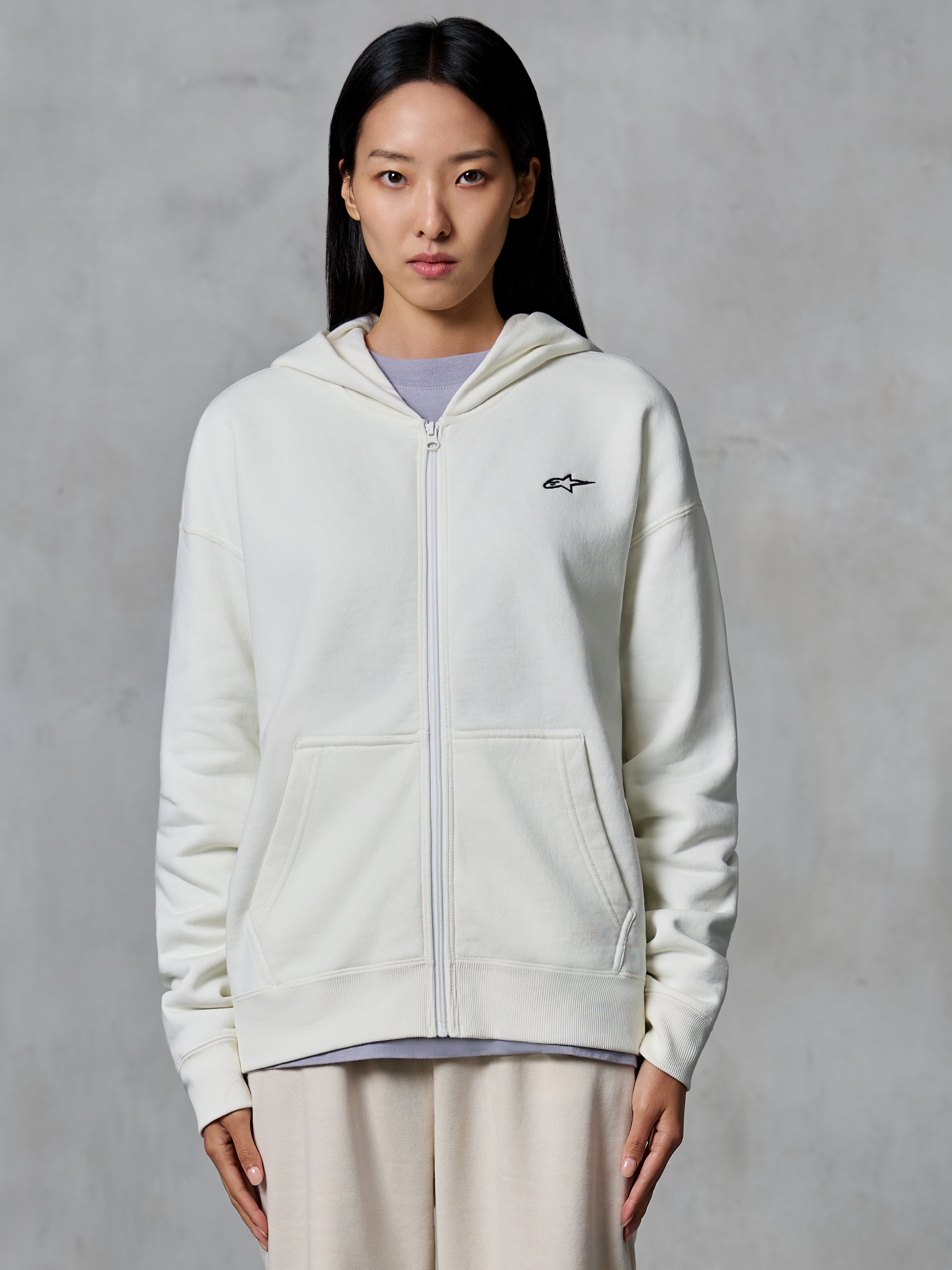 Alpinestars Essentials Ageless Zip Hoodie, Off White, felpa in pile con zip frontale indossata da una modella, con un piccolo logo Astars nero sul petto, vestibilità rilassata, abbigliamento lifestyle casual ispirato al motorsport