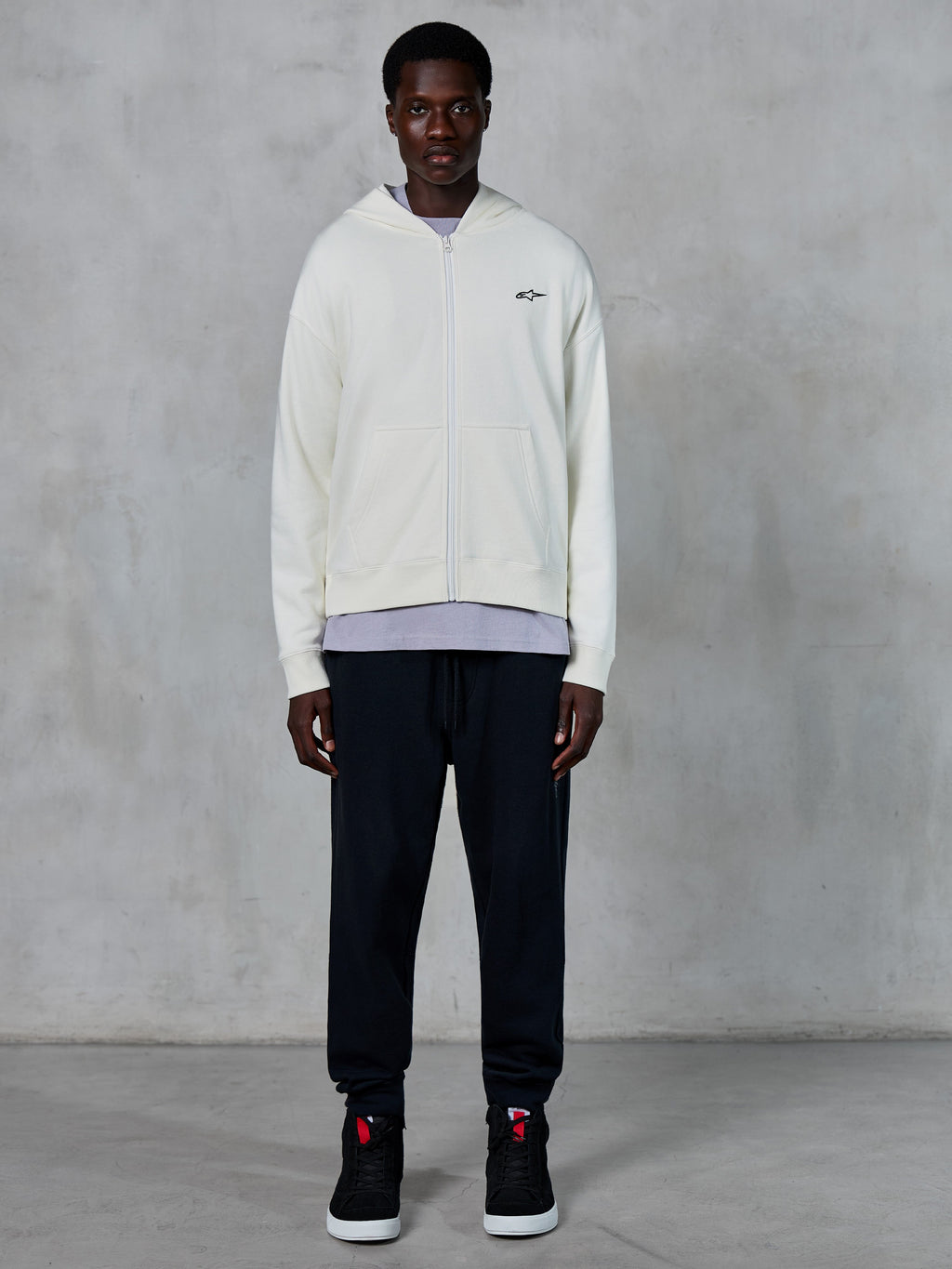 Alpinestars Essentials Ageless Zip Hoodie, felpa con zip integrale bianco panna, vestibilità rilassata con piccolo logo nero sul petto, indossata con pantaloni della tuta neri e sneakers, streetwear casual ispirato al motorsport.