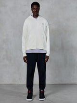 Alpinestars Essentials Ageless Zip Hoodie, felpa con zip integrale bianco panna, vestibilità rilassata con piccolo logo nero sul petto, indossata con pantaloni della tuta neri e sneakers, streetwear casual ispirato al motorsport.