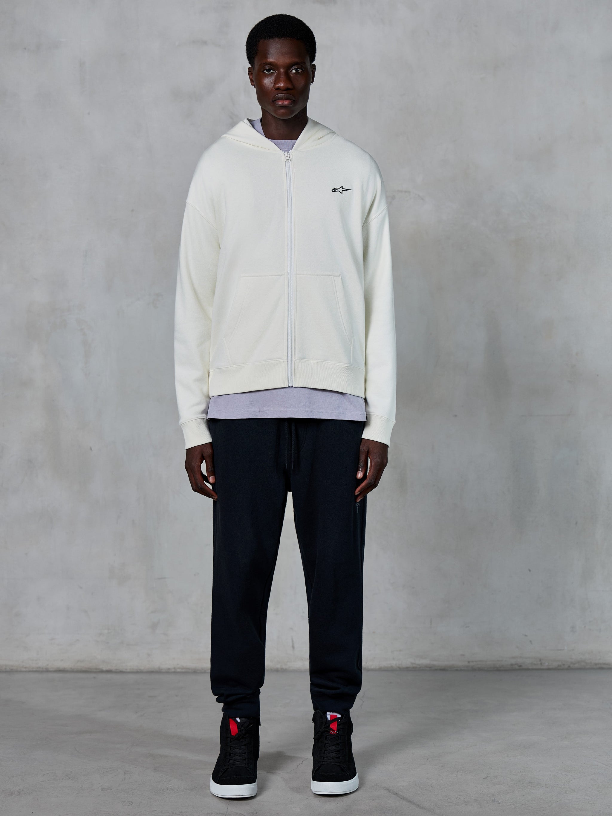 Alpinestars Essentials Ageless Zip Hoodie, felpa con zip integrale bianco panna, vestibilità rilassata con piccolo logo nero sul petto, indossata con pantaloni della tuta neri e sneakers, streetwear casual ispirato al motorsport.