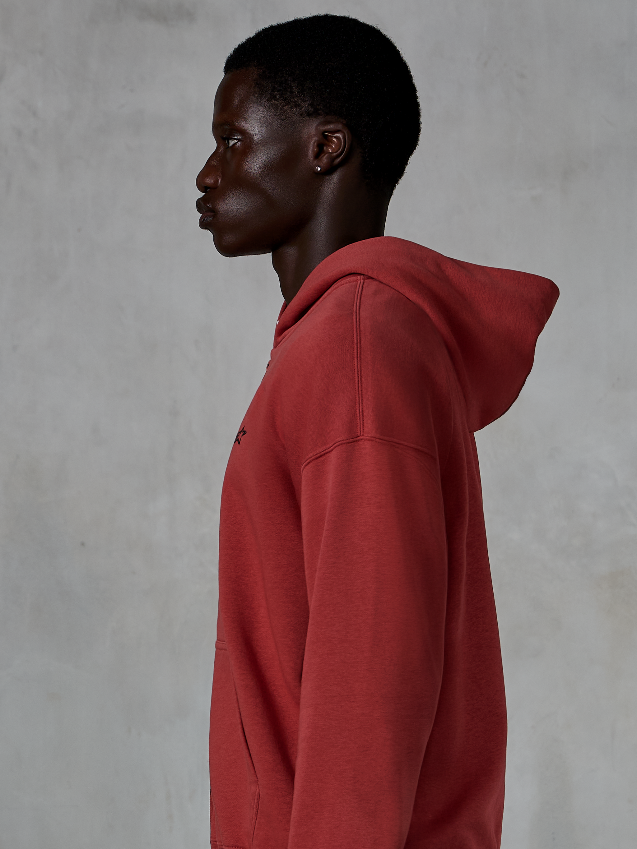 Alpinestars Essentials Ageless Zip Hoodie, rosso mattone, vista laterale di profilo di un uomo che indossa una felpa con cappuccio e zip, piccolo logo a stella nero sul petto, pile premium con lavaggio a pietra, streetwear casual ispirato al motorsport