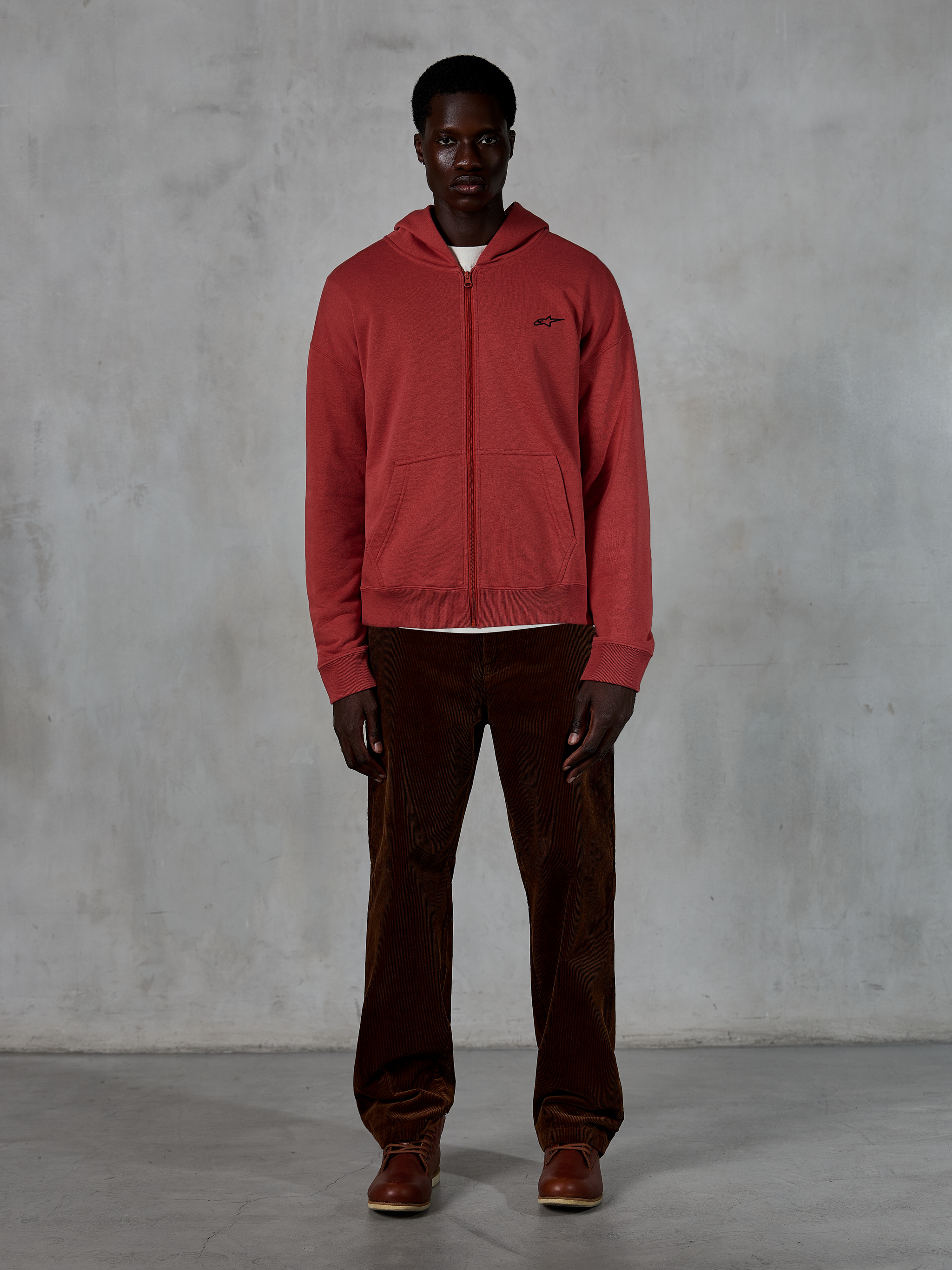 Alpinestars Essentials Ageless Zip Hoodie, felpa casual streetwear, rosso mattone con piccolo logo a stella nero, zip integrale frontale, indossata da un modello con pantaloni in velluto a coste marroni e stivali, design premium ispirato al motorsport