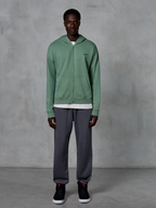 Alpinestars Essentials Ageless Zip Hoodie, felpa con cappuccio e zip, verde cactus, indossata da un modello con joggers grigio scuro e sneakers nere, caratterizzata da un piccolo logo Alpinestars nero sul petto, vestibilità rilassata, streetwear casual.