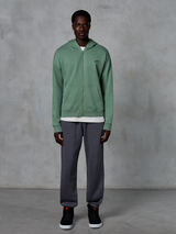 Alpinestars Essentials Ageless Zip Hoodie, felpa con cappuccio e zip, verde cactus, indossata da un modello con joggers grigio scuro e sneakers nere, caratterizzata da un piccolo logo Alpinestars nero sul petto, vestibilità rilassata, streetwear casual.