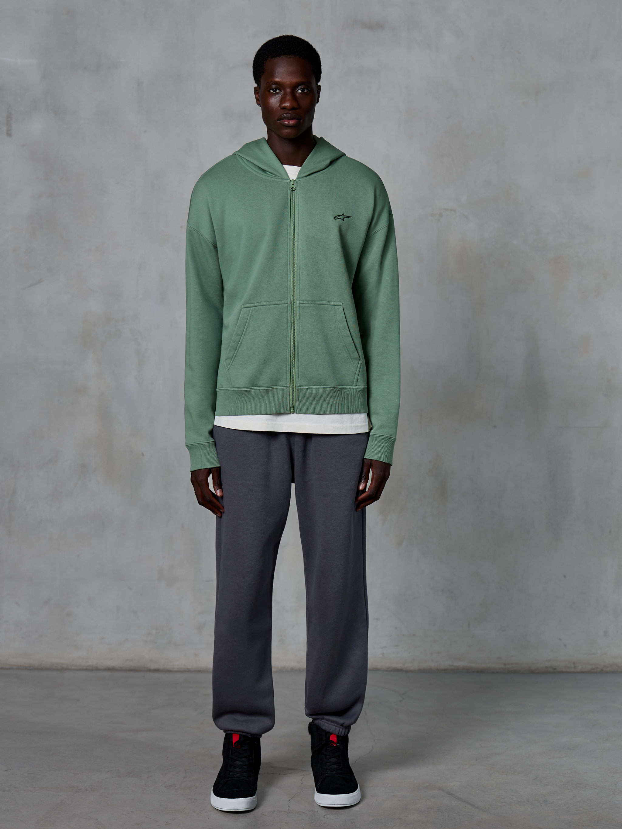 Alpinestars Essentials Ageless Zip Hoodie, felpa con cappuccio e zip, verde cactus, indossata da un modello con joggers grigio scuro e sneakers nere, caratterizzata da un piccolo logo Alpinestars nero sul petto, vestibilità rilassata, streetwear casual.