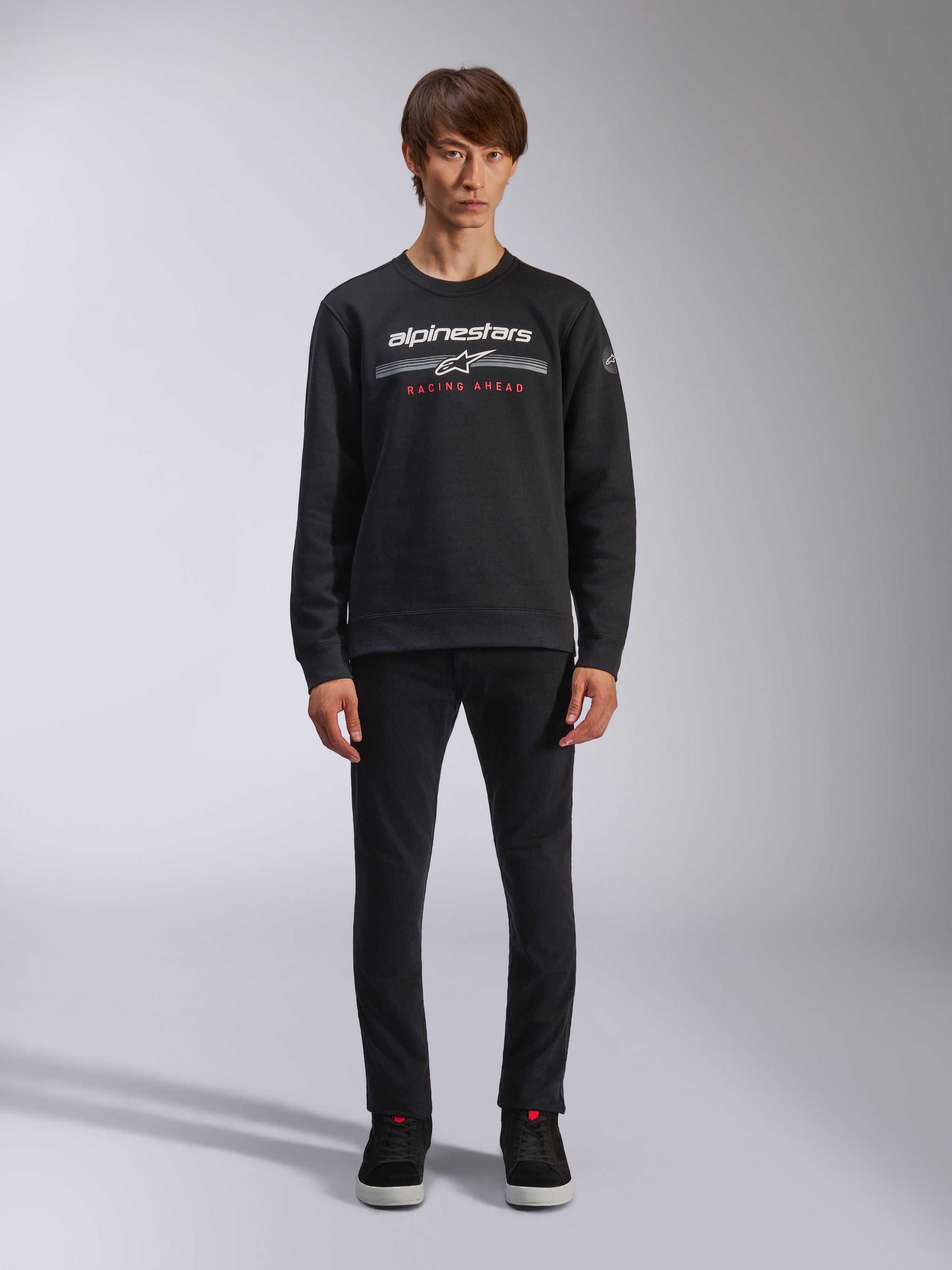 Alpinestars Bettern Crew, felpa a girocollo nera, con logo Alpinestars bianco, strisce orizzontali grigie e scritta 'RACING AHEAD' rossa, streetwear casual motorsport, indossata da un modello con pantaloni neri e sneakers