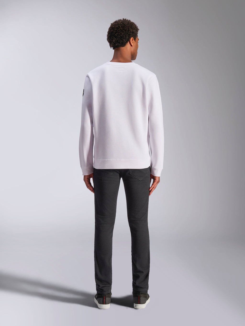 Alpinestars Bettern Crew, felpa a girocollo bianca, vista posteriore su modello, abbinata a jeans scuri e sneakers, streetwear casual ispirato al motorsport