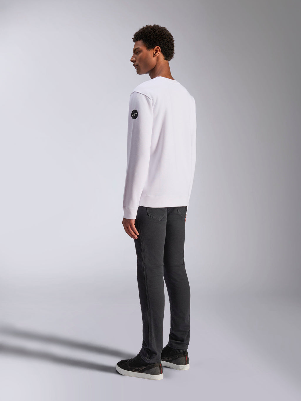 Alpinestars Bettern Crew, felpa girocollo bianca, vista posteriore e laterale, logo circolare nero sulla manica, indossata con pantaloni grigio scuro e sneakers nere, streetwear casual lifestyle motorsport
