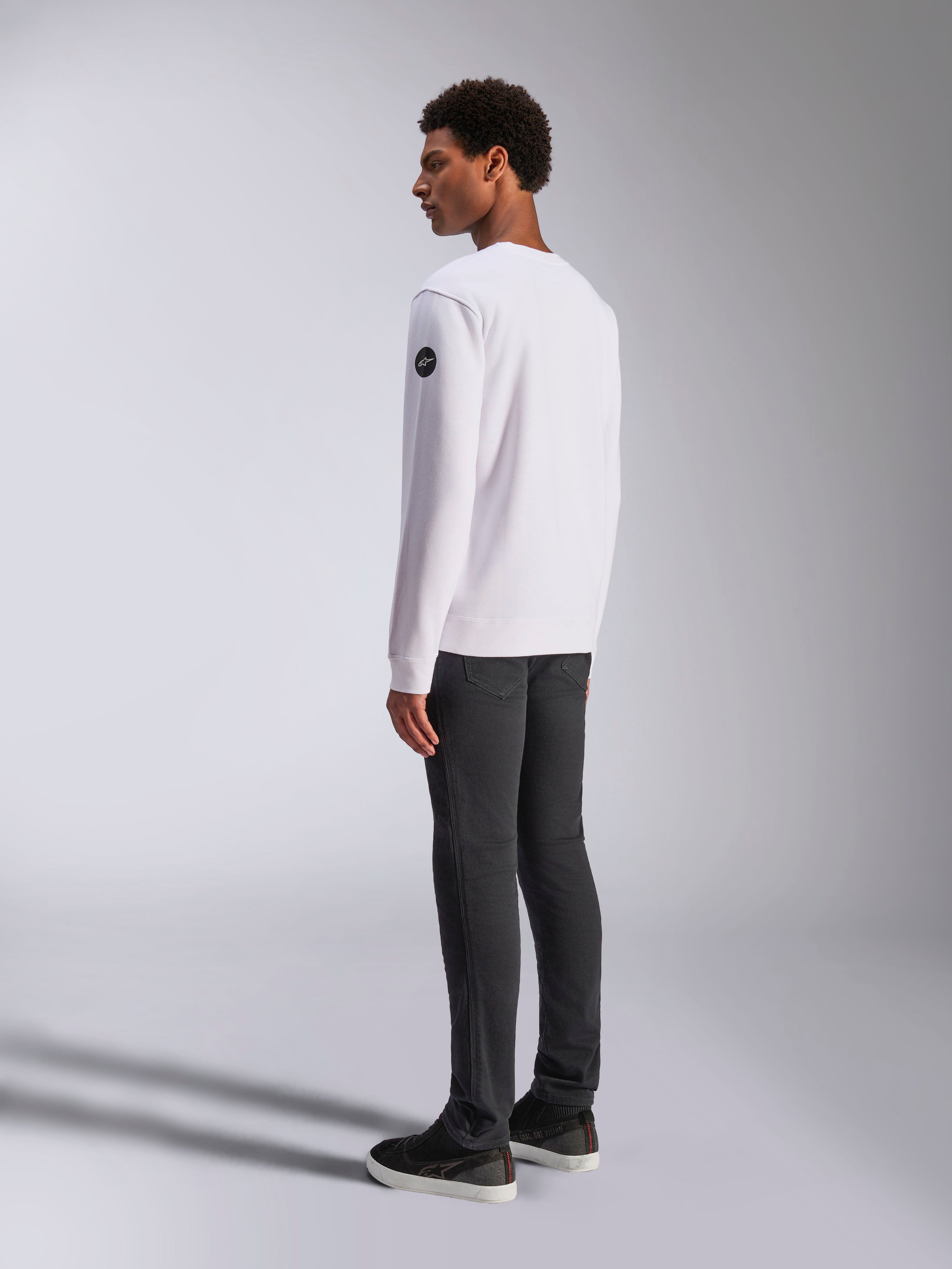 Alpinestars Bettern Crew, felpa girocollo bianca, vista posteriore e laterale, logo circolare nero sulla manica, indossata con pantaloni grigio scuro e sneakers nere, streetwear casual lifestyle motorsport