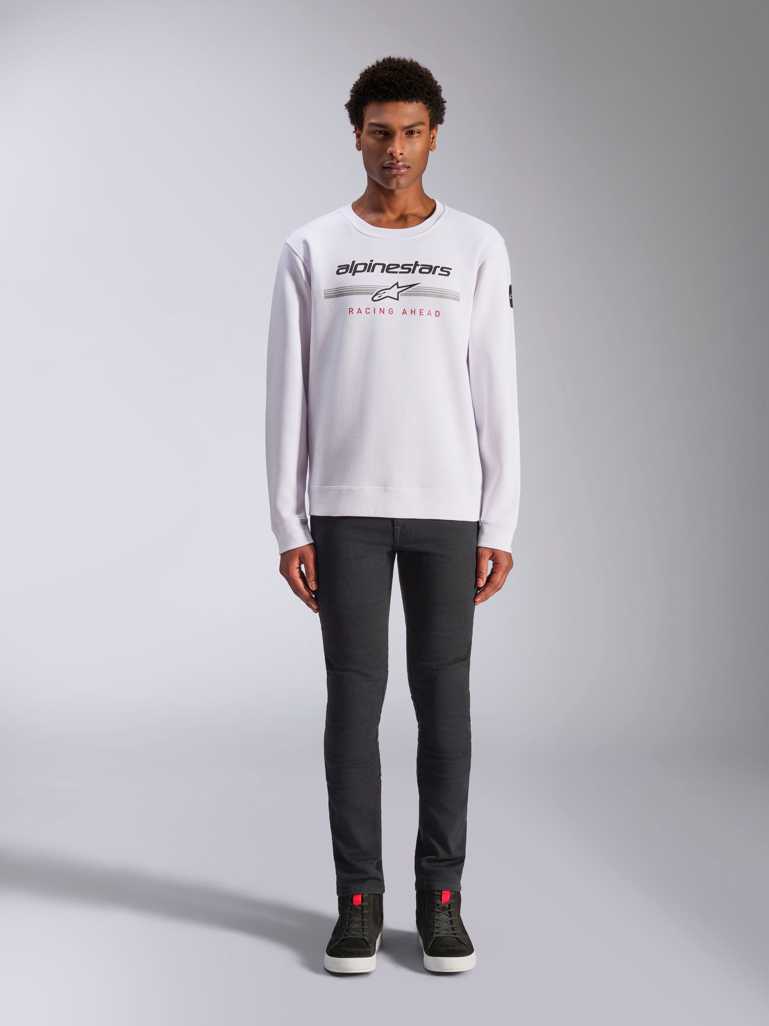 Alpinestars Bettern Crew, felpa girocollo bianca, indossata da un modello con pantaloni grigio scuro e sneakers alte nere, con marchio Alpinestars e scritta rossa 'Racing Ahead', streetwear casual ispirato al motorsport