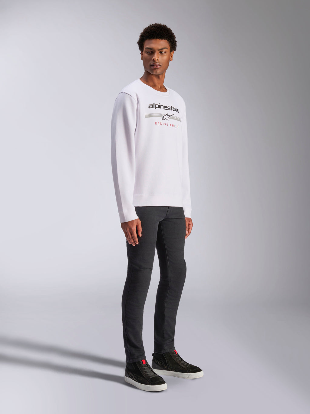 Alpinestars Bettern Crew, felpa girocollo bianca, con logo Alpinestars nero e scritta rossa 'Racing Ahead', streetwear casual motorsport, indossata da un modello con pantaloni scuri e sneakers nere