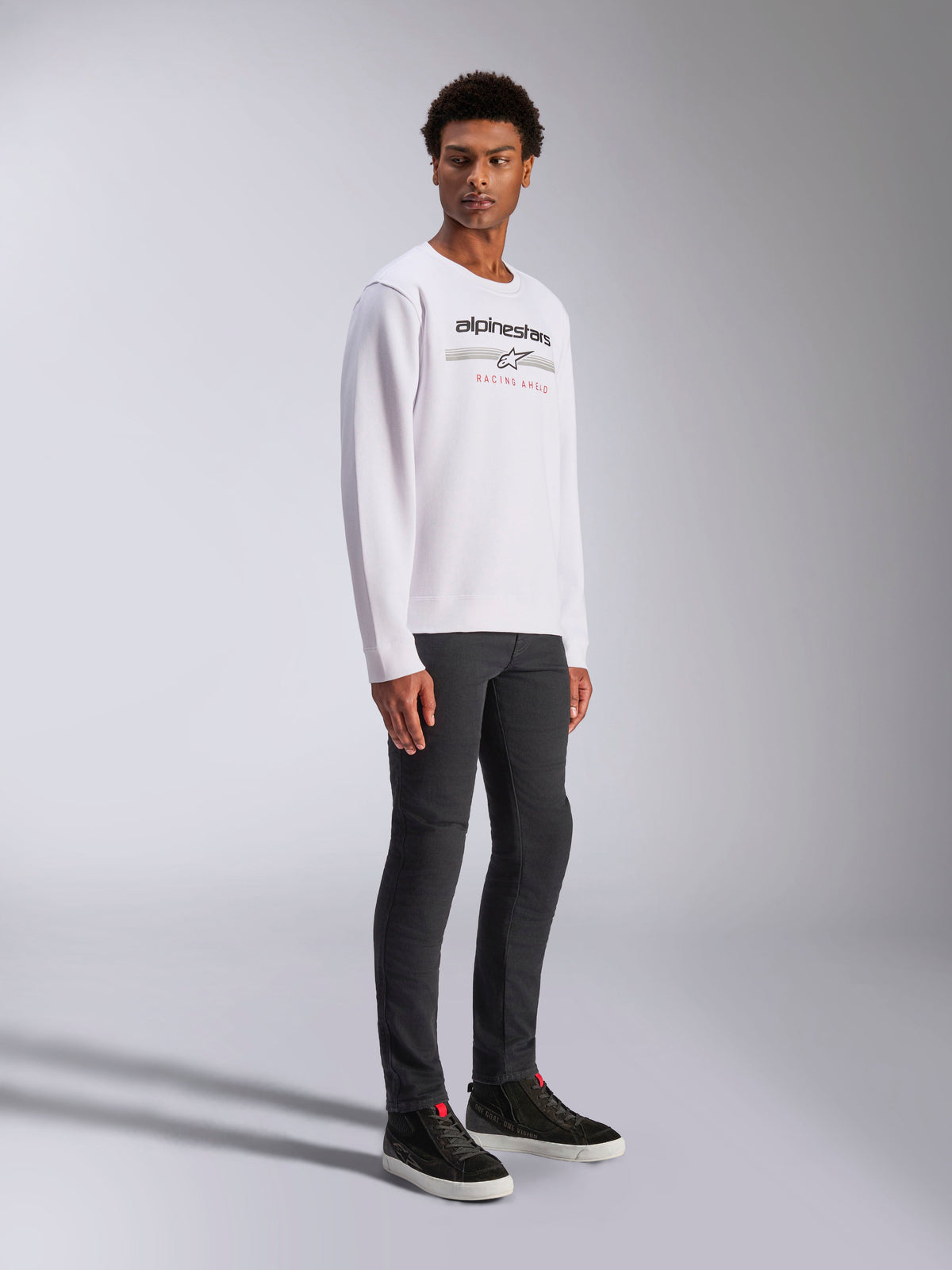 Alpinestars Bettern Crew, felpa girocollo bianca, con logo Alpinestars nero e scritta rossa 'Racing Ahead', streetwear casual motorsport, indossata da un modello con pantaloni scuri e sneakers nere