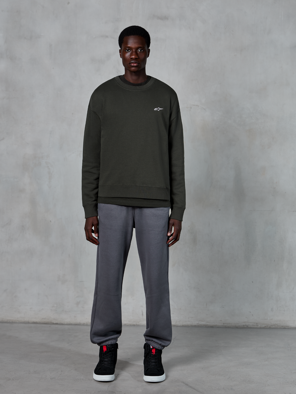 Alpinestars Essentials Ageless Crew, felpa girocollo verde scuro con un piccolo logo bianco sul petto, indossata da un modello con pantaloni della tuta grigi e sneakers alte nere, abbigliamento lifestyle casual