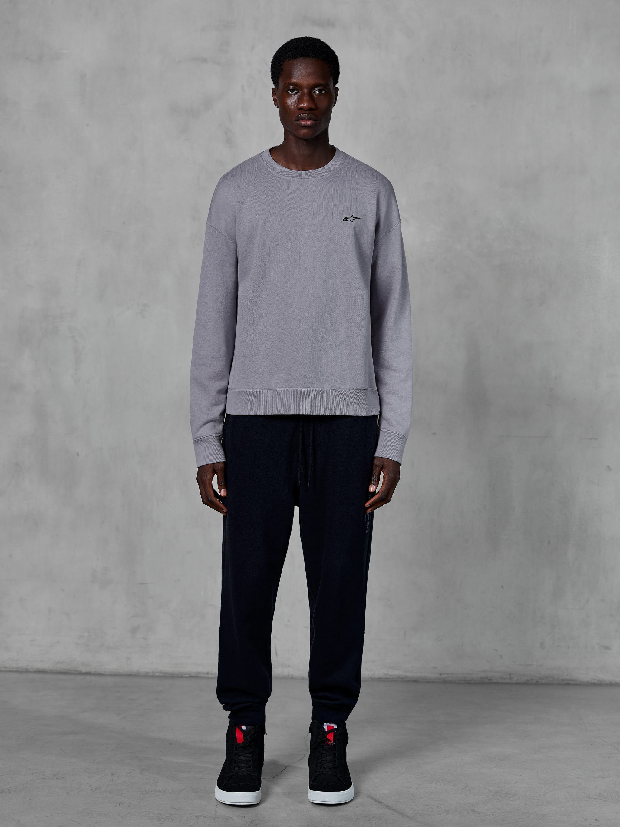 Alpinestars Essentials Ageless Crew, felpa girocollo argento, indossata da un modello con pantaloni jogger neri e sneakers nere, con un piccolo logo Alpinestars nero sul petto, streetwear casual ispirato al motorsport