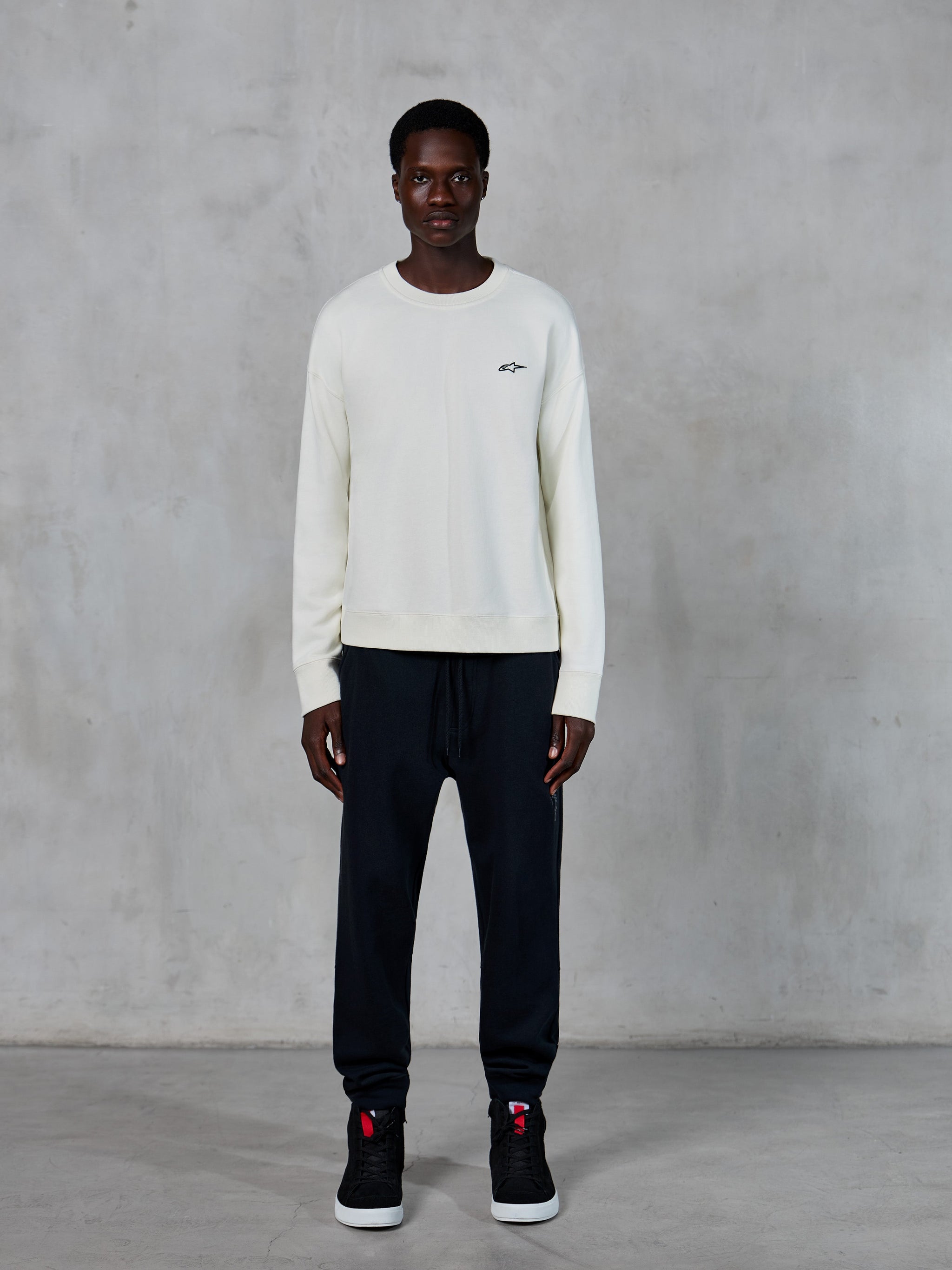 Alpinestars Essentials Ageless Crew, felpa girocollo, Off White con piccolo logo a stella nero, indossata da un modello con joggers neri, streetwear casual ispirato al motorsport