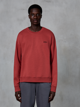 Alpinestars Essentials Ageless Crew, felpa girocollo, rosso mattone, piccolo logo nero sul petto, indossata da un modello con pantaloni grigi, streetwear casual in pile morbido premium