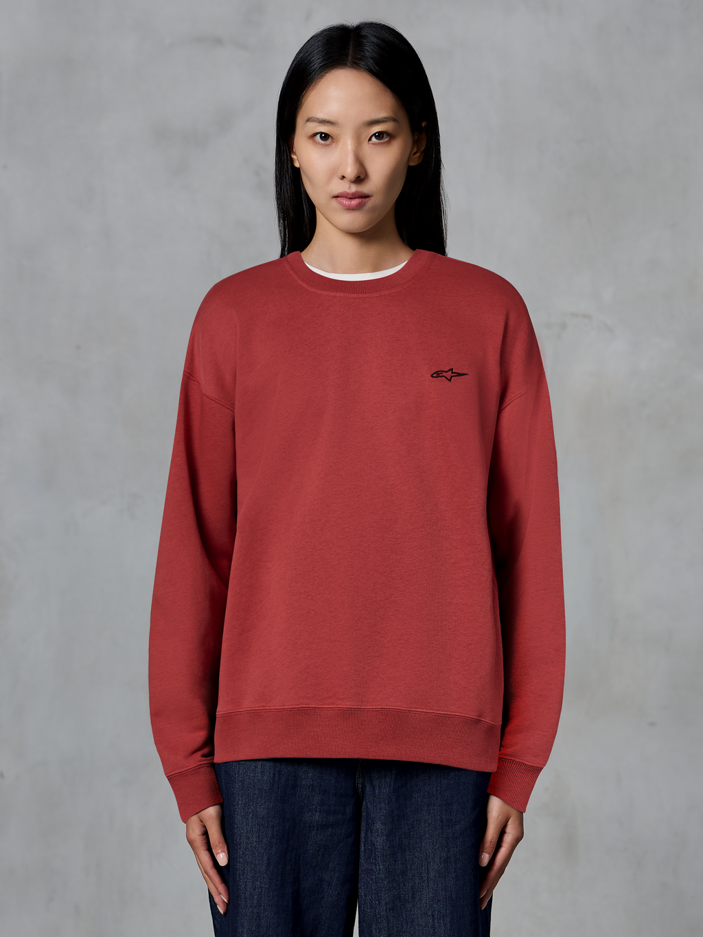 Alpinestars Essentials Ageless Crew, felpa girocollo rosso mattone, piccolo logo nero sul petto, pile premium, indossata da una modella, streetwear casual ispirato al motorsport