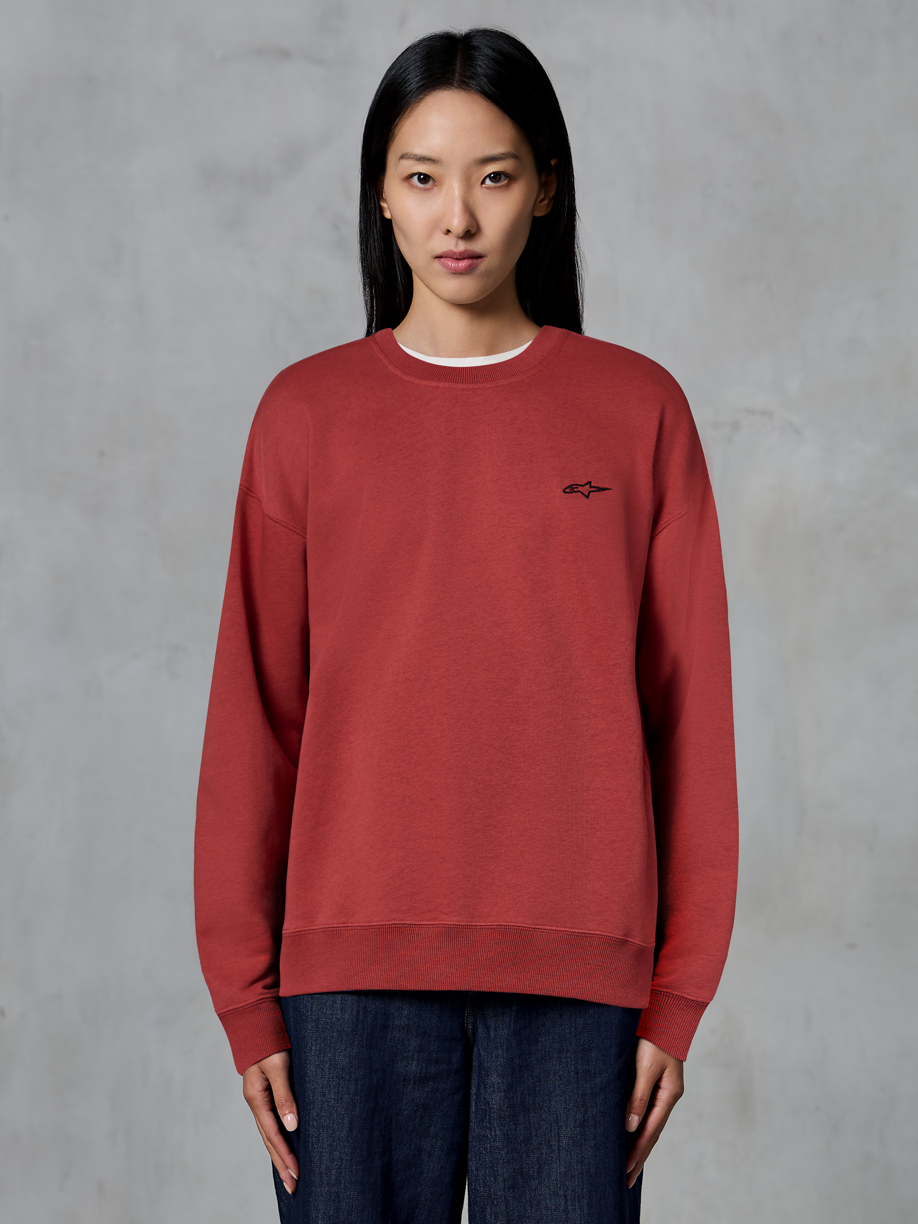 Alpinestars Essentials Ageless Crew, felpa girocollo rosso mattone, piccolo logo nero sul petto, pile premium, indossata da una modella, streetwear casual ispirato al motorsport