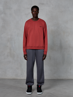 Alpinestars Essentials Ageless Crew, felpa girocollo rosso mattone, indossata con pantaloni della tuta grigi e sneakers nere, con un piccolo logo nero sul petto, streetwear casual ispirato al motorsport