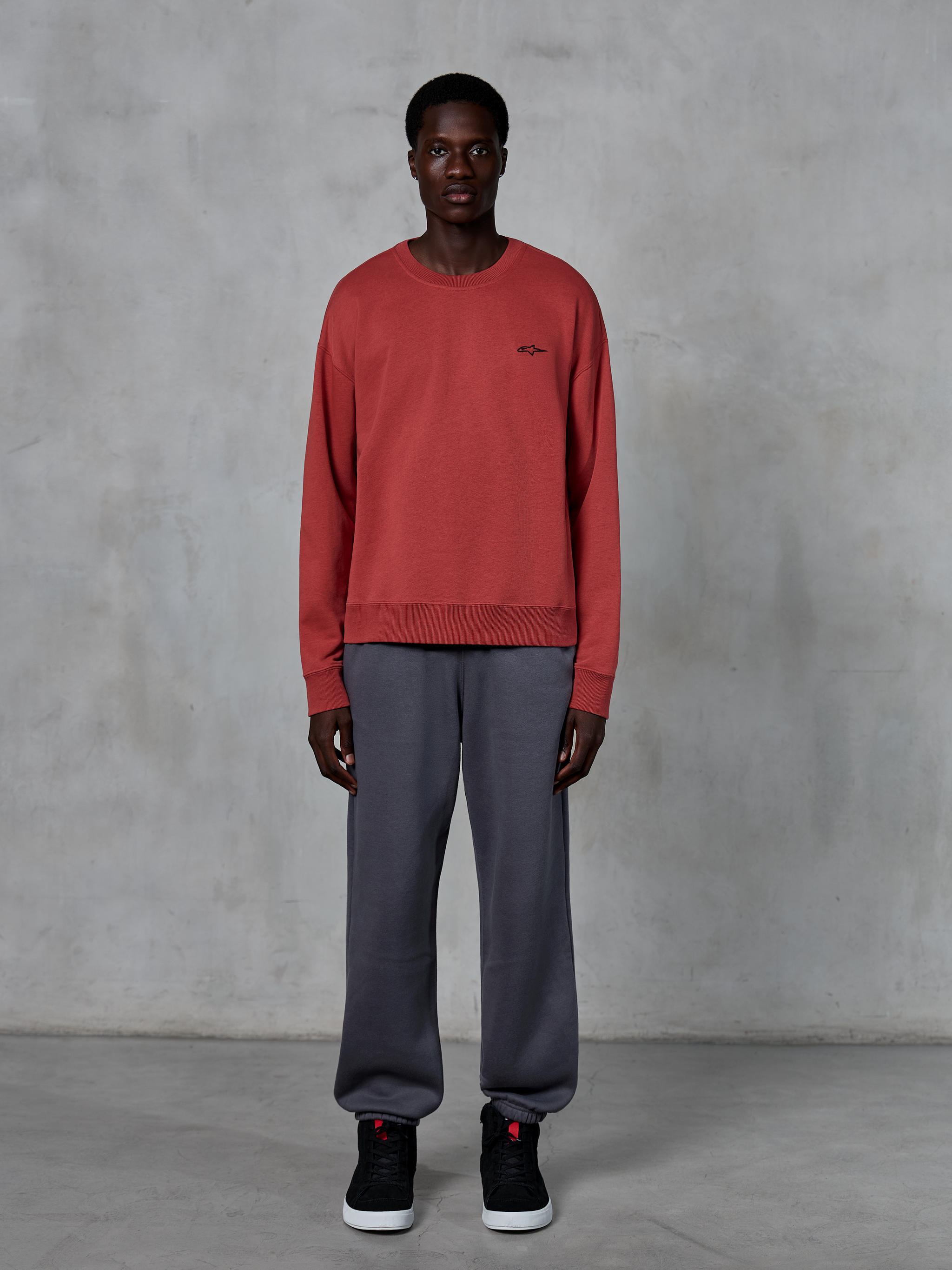 Alpinestars Essentials Ageless Crew, felpa girocollo rosso mattone, indossata con pantaloni della tuta grigi e sneakers nere, con un piccolo logo nero sul petto, streetwear casual ispirato al motorsport