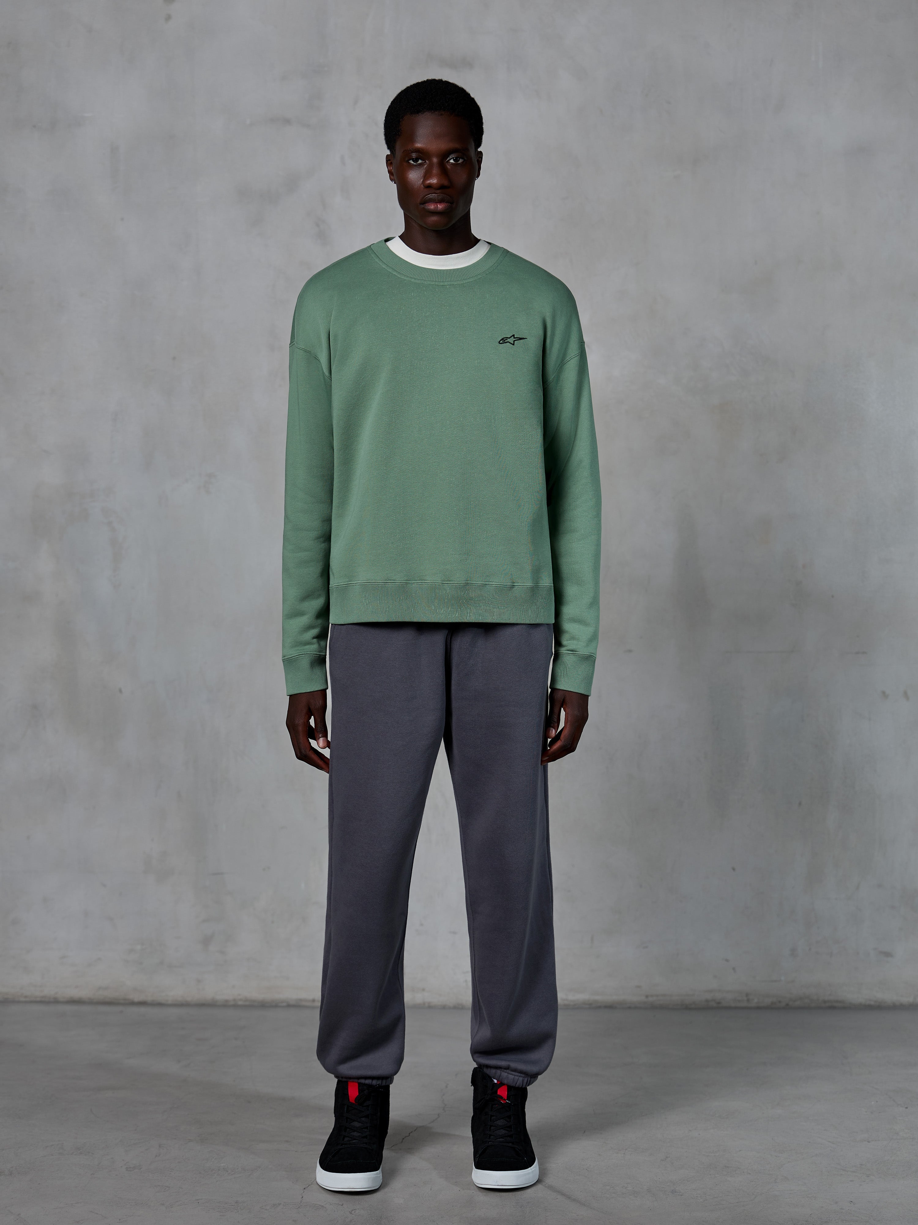 Alpinestars Essentials Ageless Crew, felpa girocollo in verde Cactus, con un piccolo logo nero sul petto, indossata da un modello con pantaloni della tuta grigi e sneakers nere, streetwear casual ispirato al motorsport