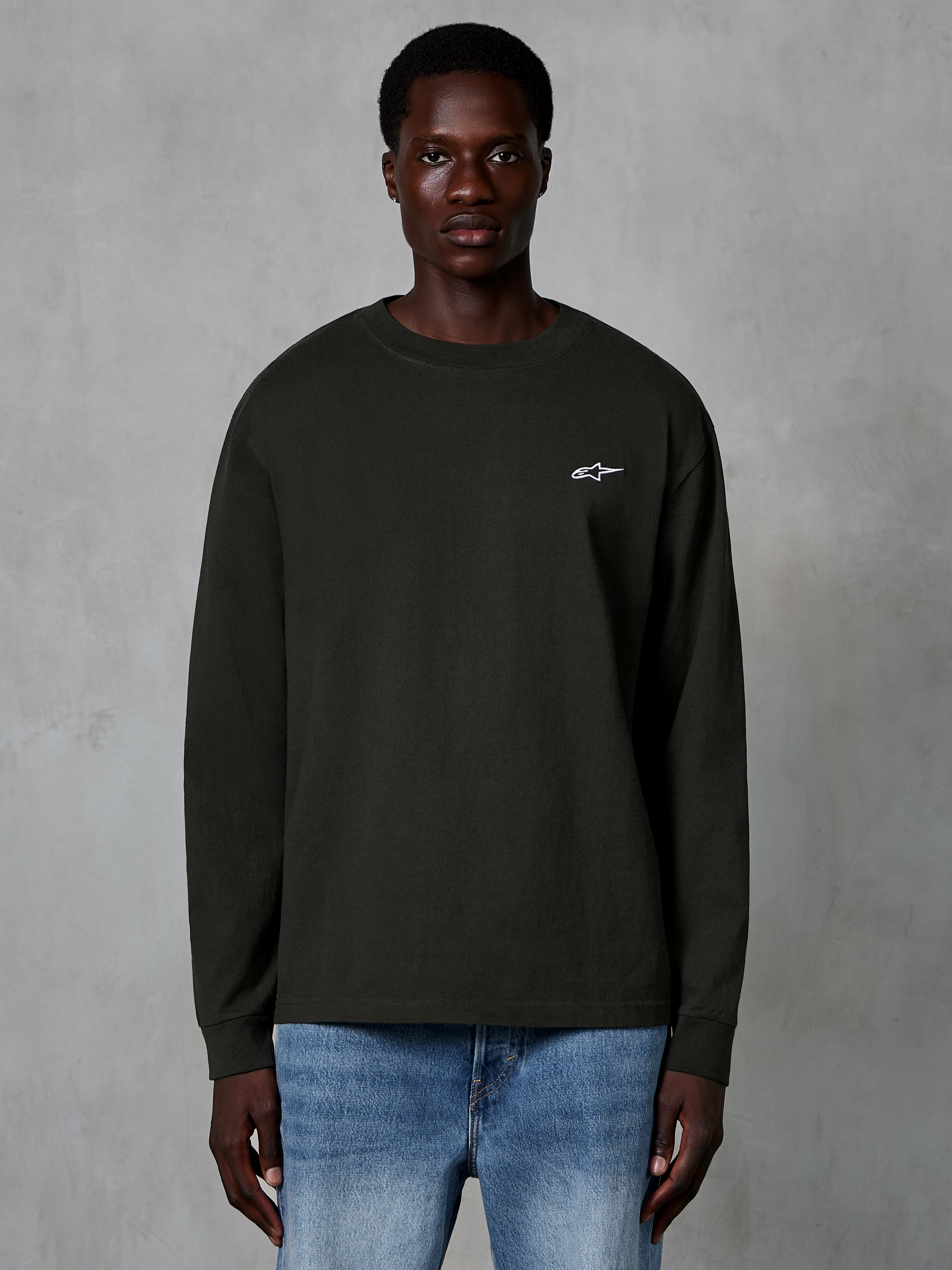 Alpinestars Essentials Ageless LS Tee, maglietta a maniche lunghe, Off Black, con un piccolo logo Alpinestars bianco sul petto, indossata da un modello con jeans blu, streetwear casual in cotone organico