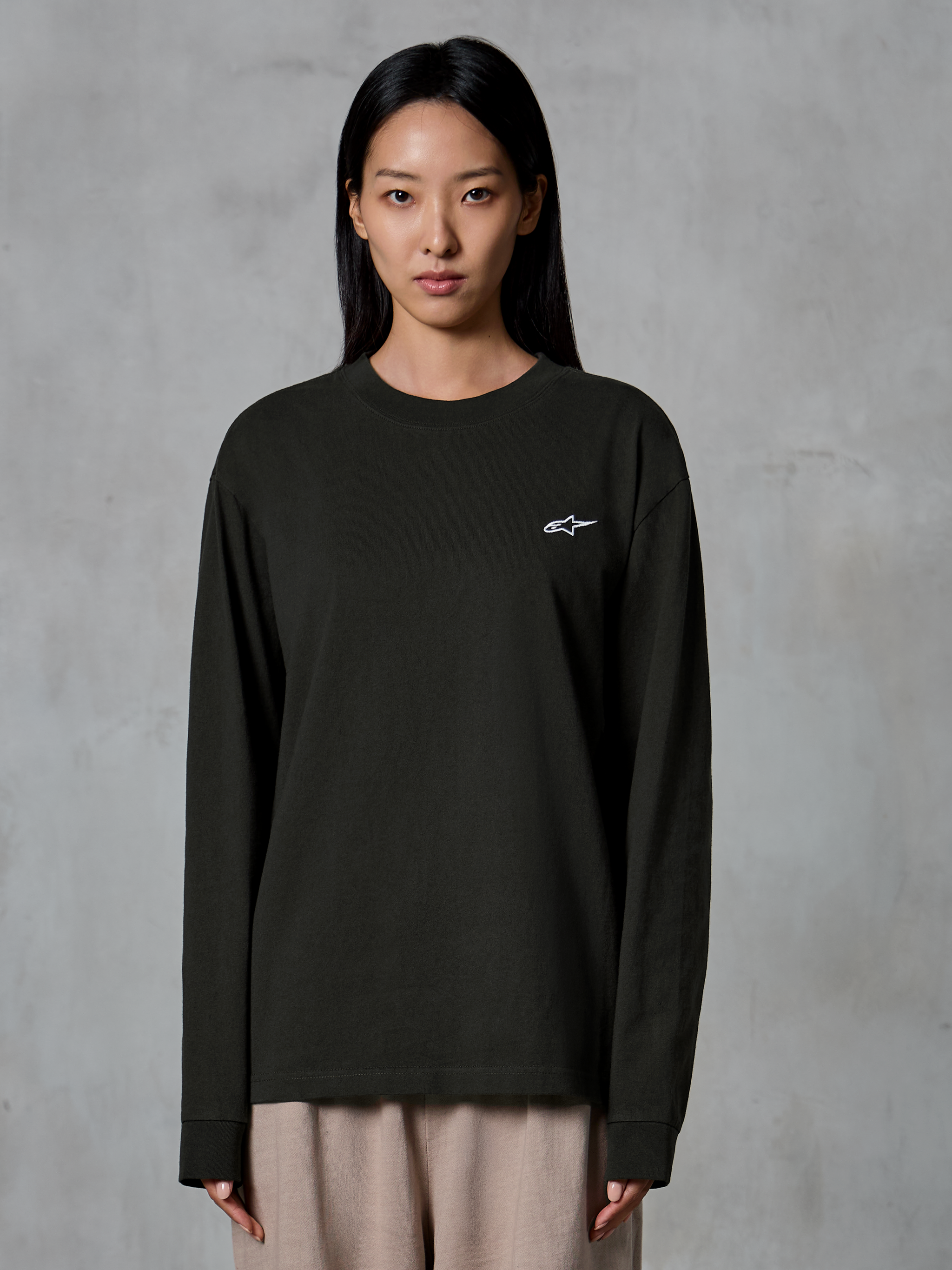 Modella che indossa Alpinestars Essentials Ageless LS Tee, t-shirt a maniche lunghe, Off Black, con un piccolo logo Alpinestars bianco sul petto, abbinata a pantaloni beige, stile casual lifestyle streetwear