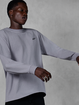 Alpinestars Essentials Ageless LS Tee, maglia a maniche lunghe, argento, piccolo logo nero sul petto, streetwear casual ispirato al motorsport, indossata da un modello su uno sfondo grigio testurizzato