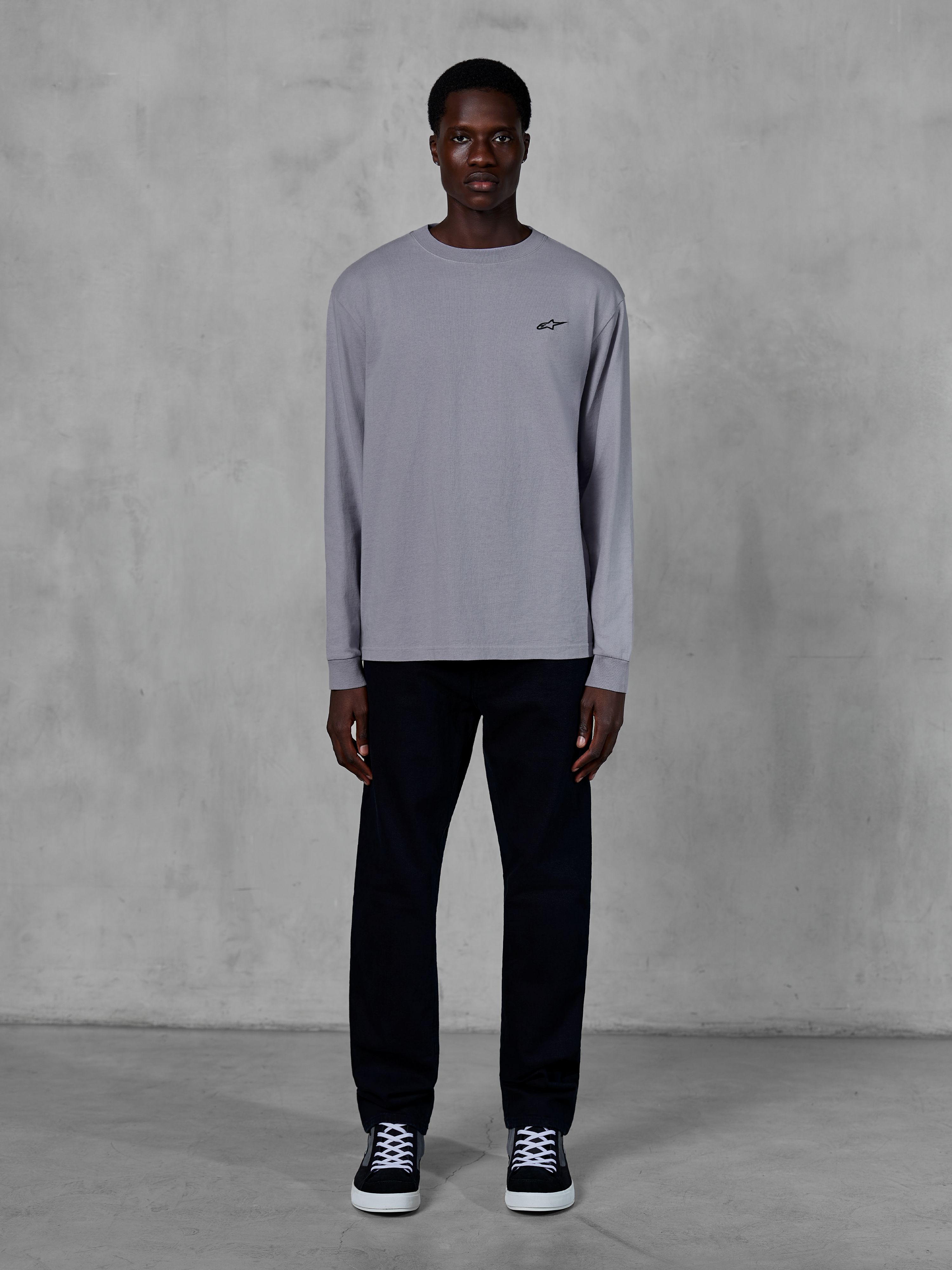 Alpinestars Essentials Ageless LS Tee, maglia a maniche lunghe, color argento, piccolo logo Alpinestars nero sul petto, jersey di cotone organico con finitura stone wash, indossata da un modello con pantaloni neri e sneakers, streetwear casual