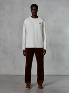 Alpinestars Essentials Ageless LS Tee, t-shirt a maniche lunghe, Off-White, indossata da un modello con pantaloni in velluto a coste marroni e stivali in pelle marrone, caratterizzata da un piccolo logo nero sul petto, vestibilità rilassata contemporanea, stile casual lifestyle