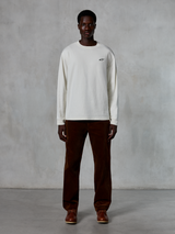 Alpinestars Essentials Ageless LS Tee, t-shirt a maniche lunghe, Off-White, indossata da un modello con pantaloni in velluto a coste marroni e stivali in pelle marrone, caratterizzata da un piccolo logo nero sul petto, vestibilità rilassata contemporanea, stile casual lifestyle