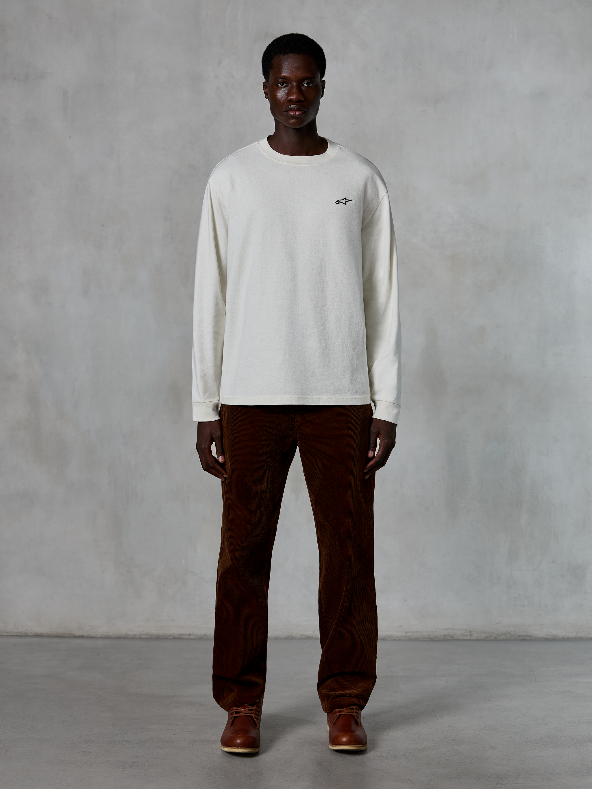 Alpinestars Essentials Ageless LS Tee, t-shirt a maniche lunghe, Off-White, indossata da un modello con pantaloni in velluto a coste marroni e stivali in pelle marrone, caratterizzata da un piccolo logo nero sul petto, vestibilità rilassata contemporanea, stile casual lifestyle
