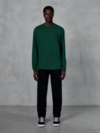 Alpinestars Essentials Ageless LS Tee, maglia a maniche lunghe, verde pino, con un piccolo logo nero sul petto, indossata da un modello con pantaloni neri e sneakers, stile streetwear casual