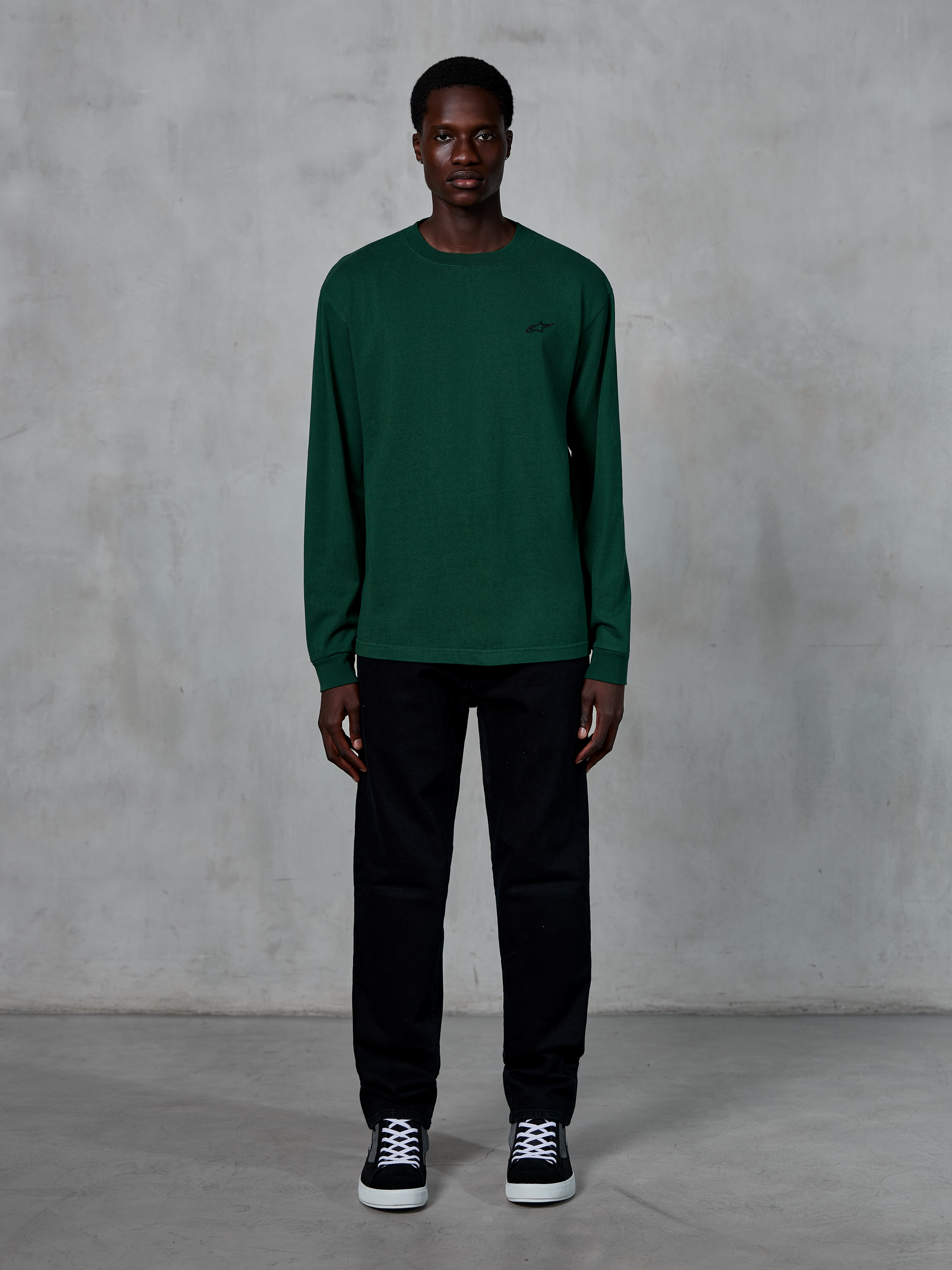 Alpinestars Essentials Ageless LS Tee, maglia a maniche lunghe, verde pino, con un piccolo logo nero sul petto, indossata da un modello con pantaloni neri e sneakers, stile streetwear casual