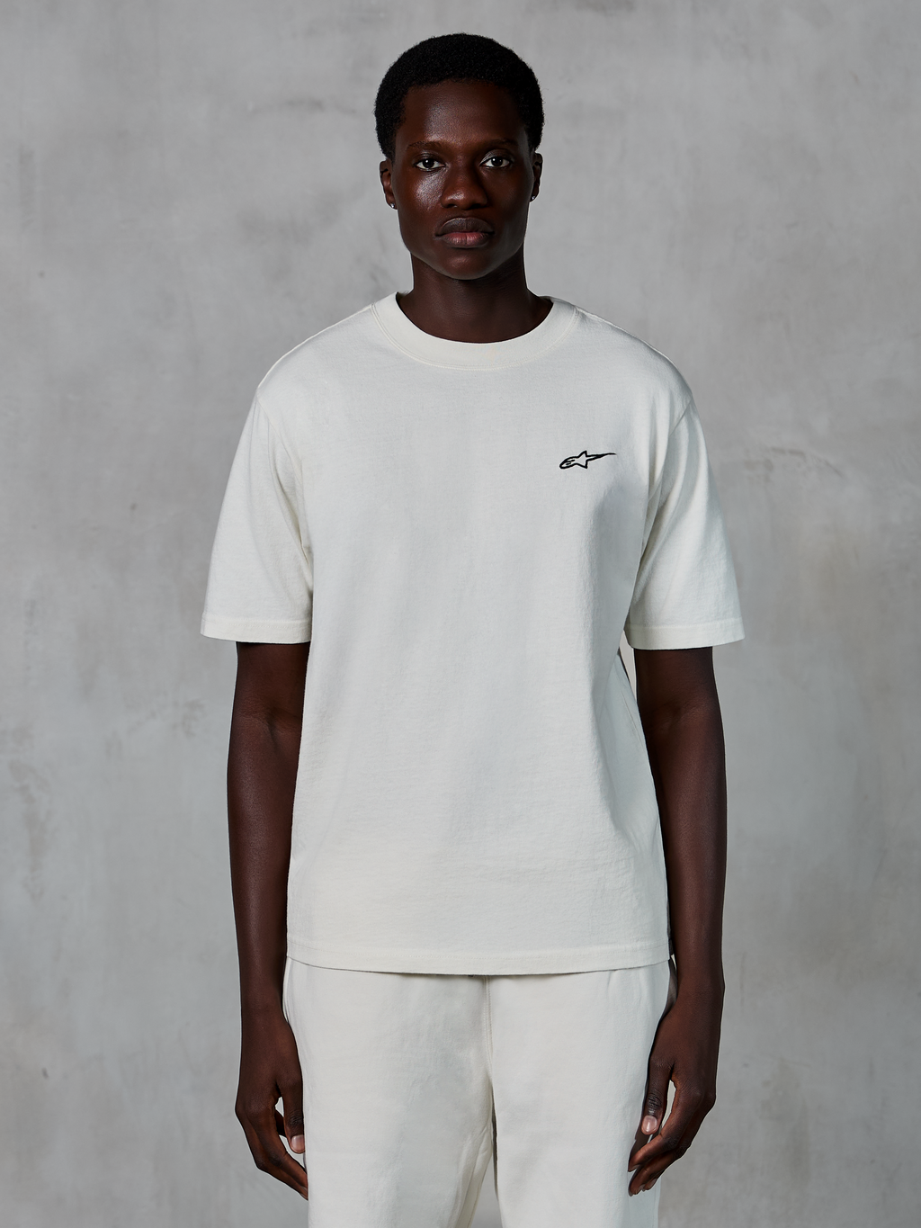 Alpinestars Essentials Ageless SS Tee, t-shirt a maniche corte, off white, indossata da un modello, con un piccolo logo Alpinestars nero sul petto, vestibilità rilassata, jersey di cotone organico, streetwear casual motorsport