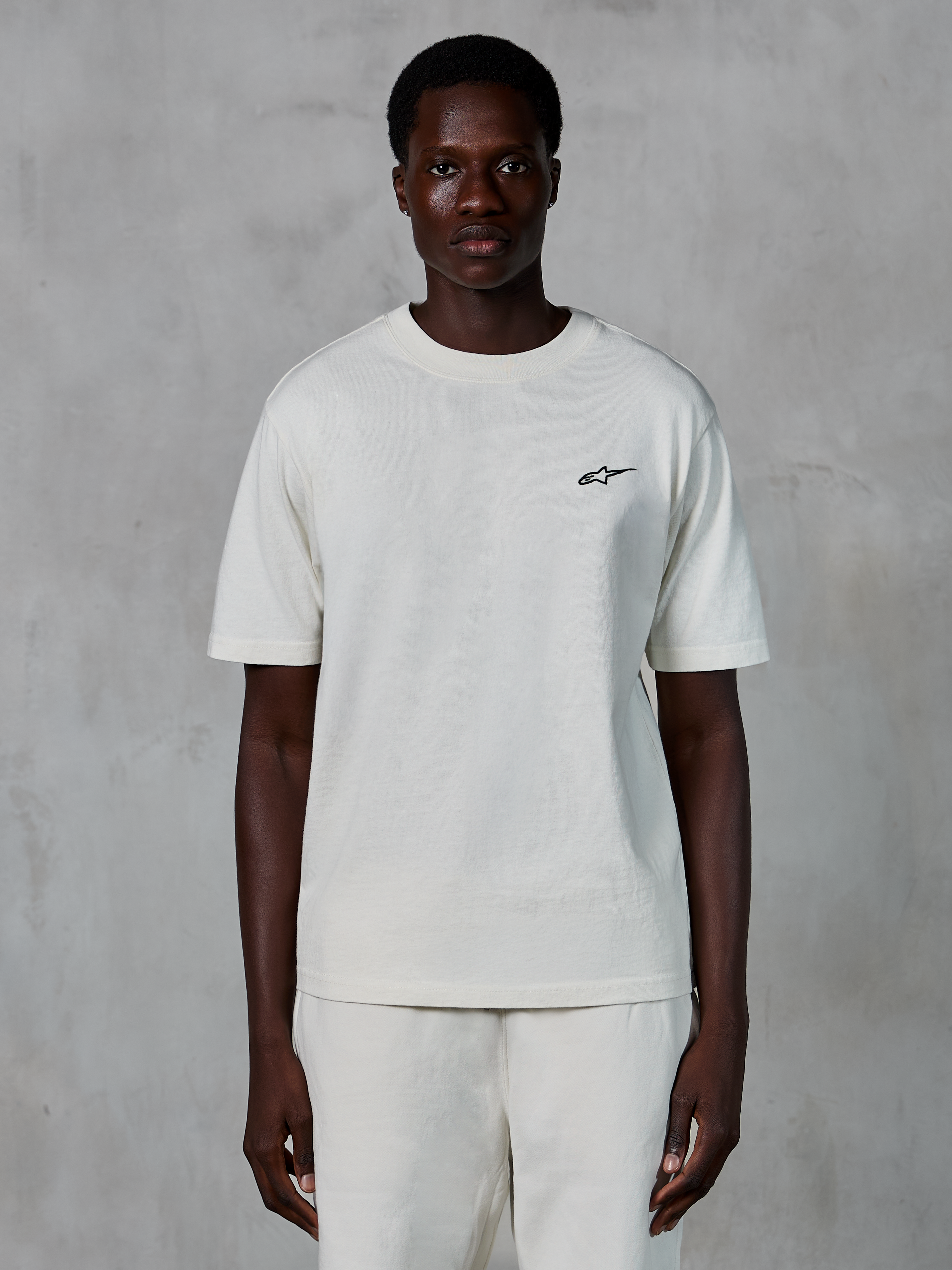 Alpinestars Essentials Ageless SS Tee, t-shirt a maniche corte, off white, indossata da un modello, con un piccolo logo Alpinestars nero sul petto, vestibilità rilassata, jersey di cotone organico, streetwear casual motorsport