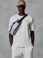 Alpinestars Essentials Ageless SS Tee, t-shirt a maniche corte, Off White, indossata con pantaloncini coordinati e un cappellino beige, con un piccolo logo nero sul petto e una borsa a tracolla grigia, streetwear casual ispirato al motorsport