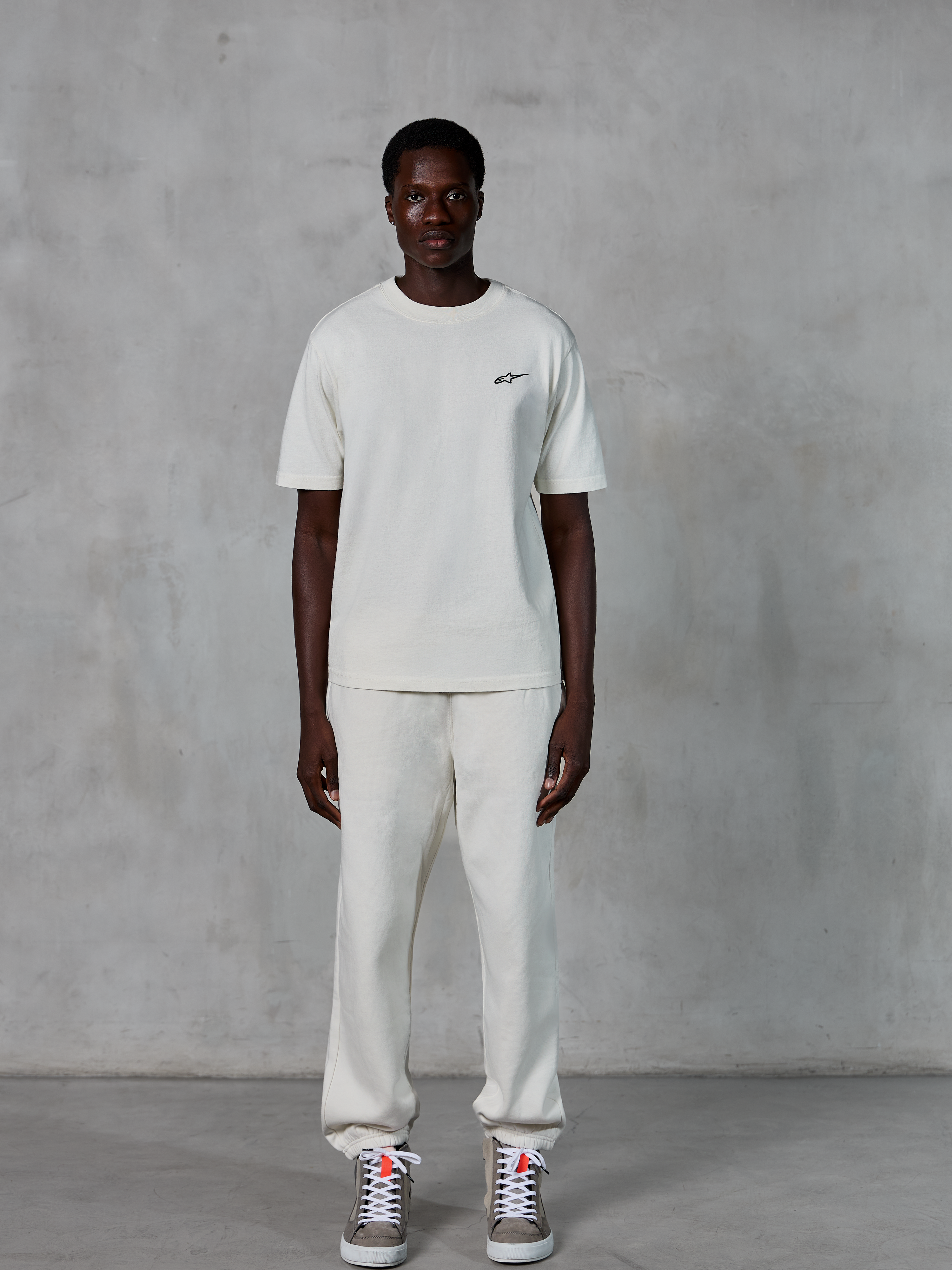 Alpinestars Essentials Ageless SS Tee, maglietta a maniche corte, bianco panna, con un piccolo logo Alpinestars nero sul petto, abbinata a pantaloni sportivi coordinati e sneakers per un look lifestyle rilassato