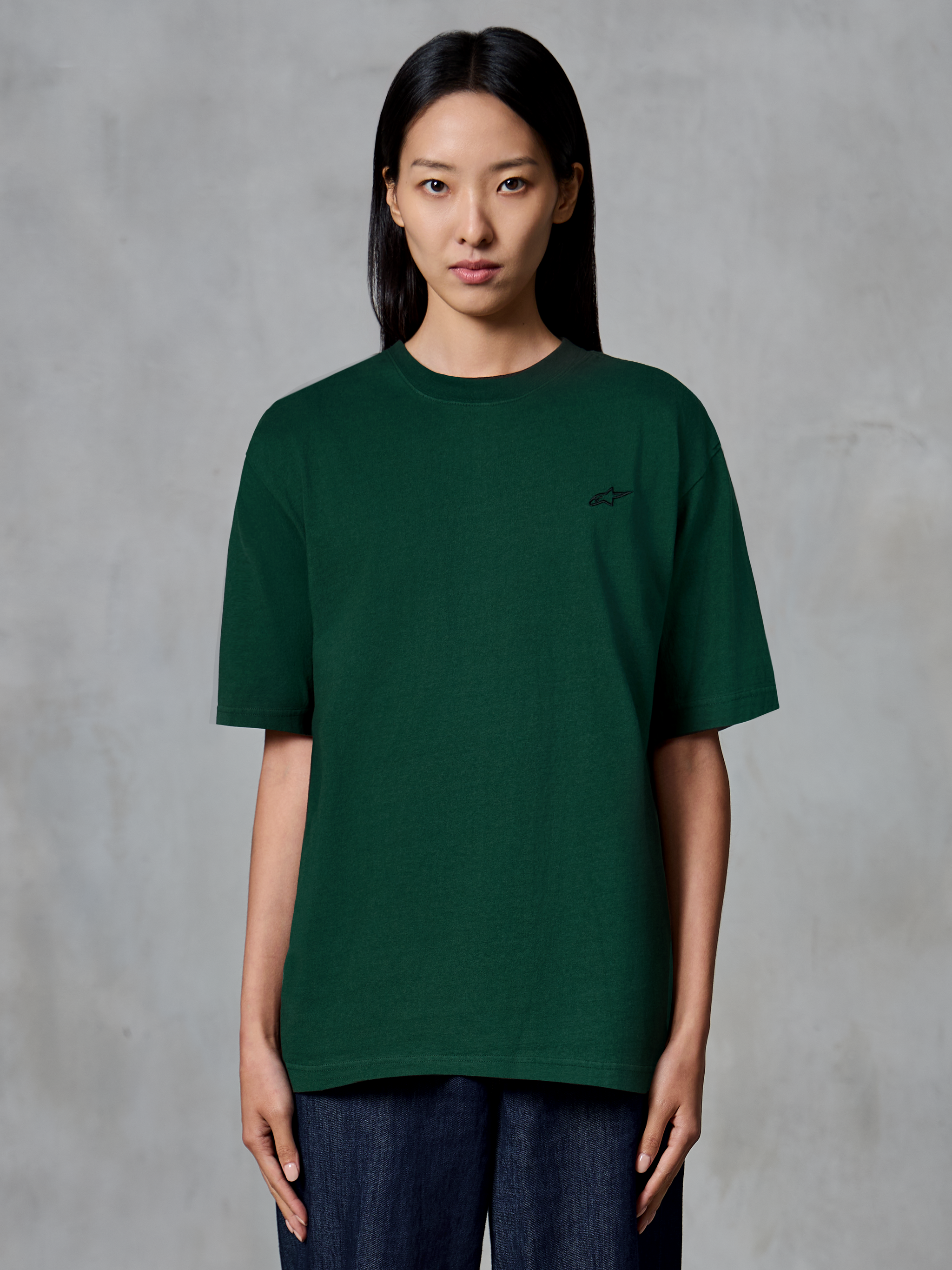 Alpinestars Essentials Ageless SS Tee, t-shirt a maniche corte, verde pino, indossata da una modella, con un piccolo logo nero sul petto, vestibilità rilassata, streetwear casual in cotone biologico